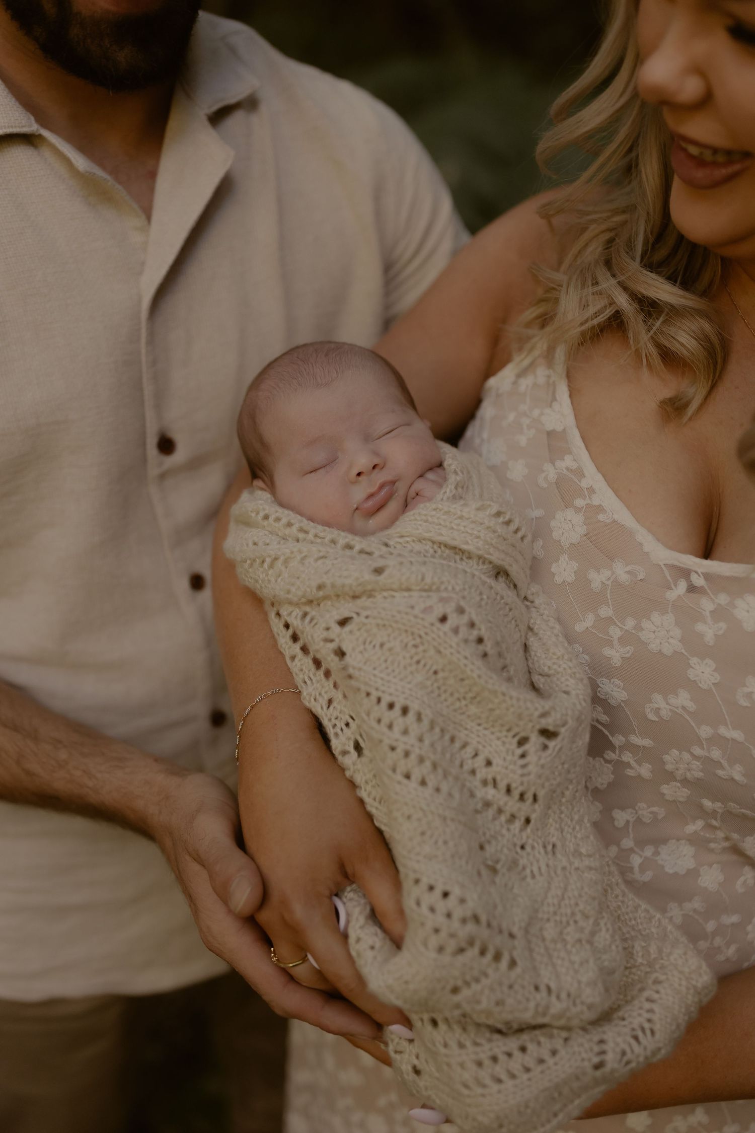 A sleeping newborn is swaddled in a cream knit blanket, cradled in mothers arms with father gently supporting.