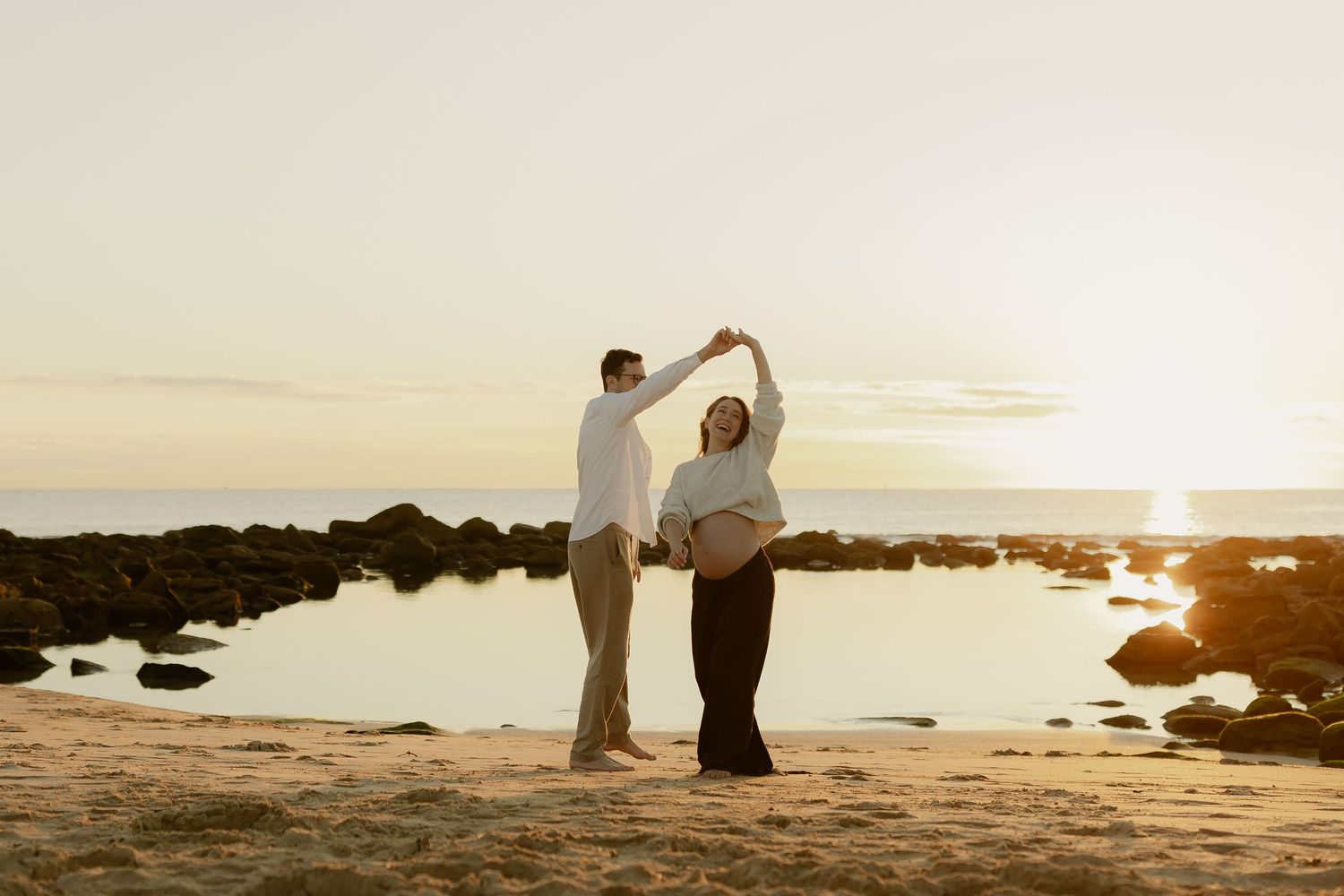 Pregnant couple dancing on a sandy beach at golden sunset, holding hands with ocean and rocks in background.