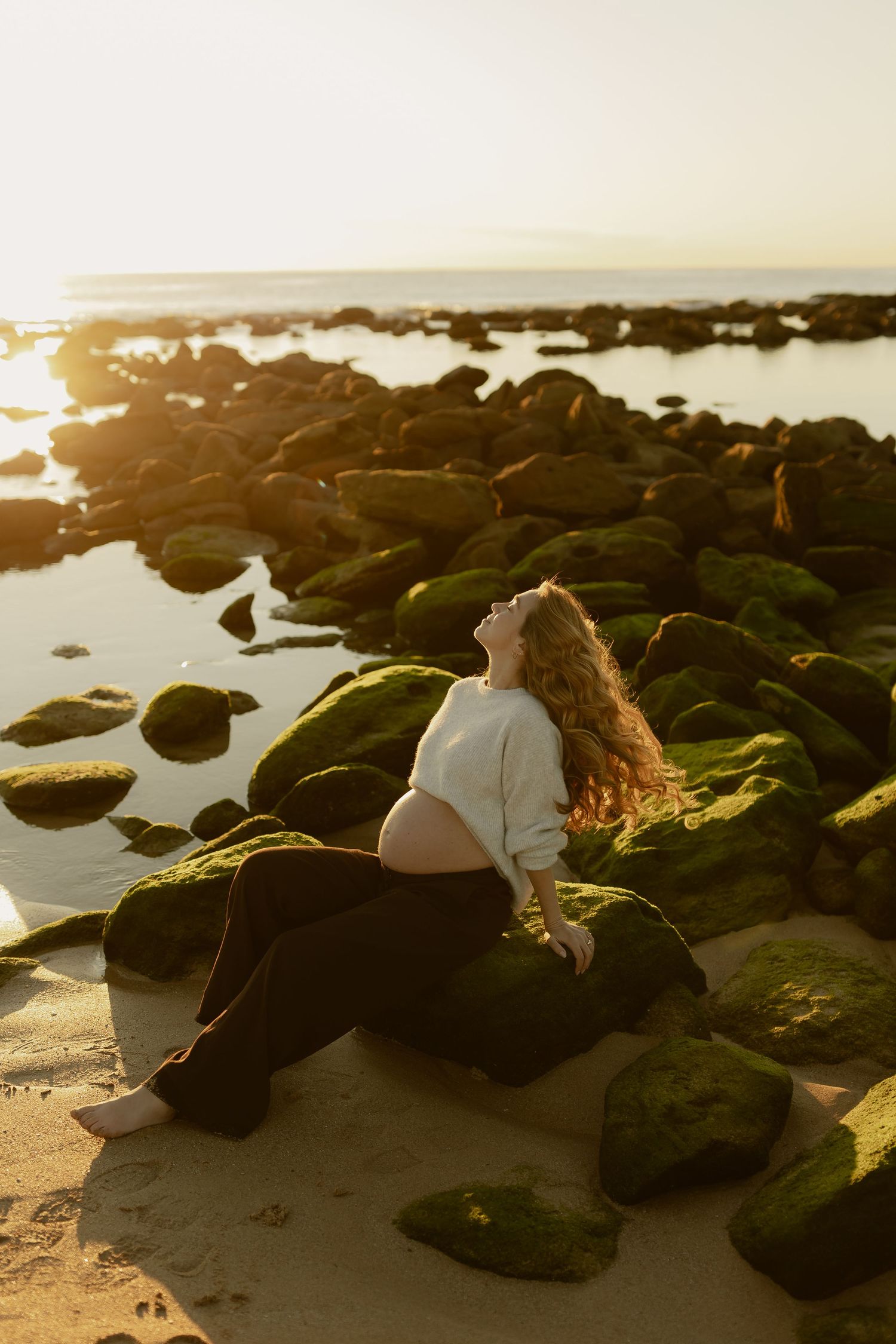 Pregnant woman sitting on moss-covered rocks at golden hour beach sunset, wearing white crop top and black pants.