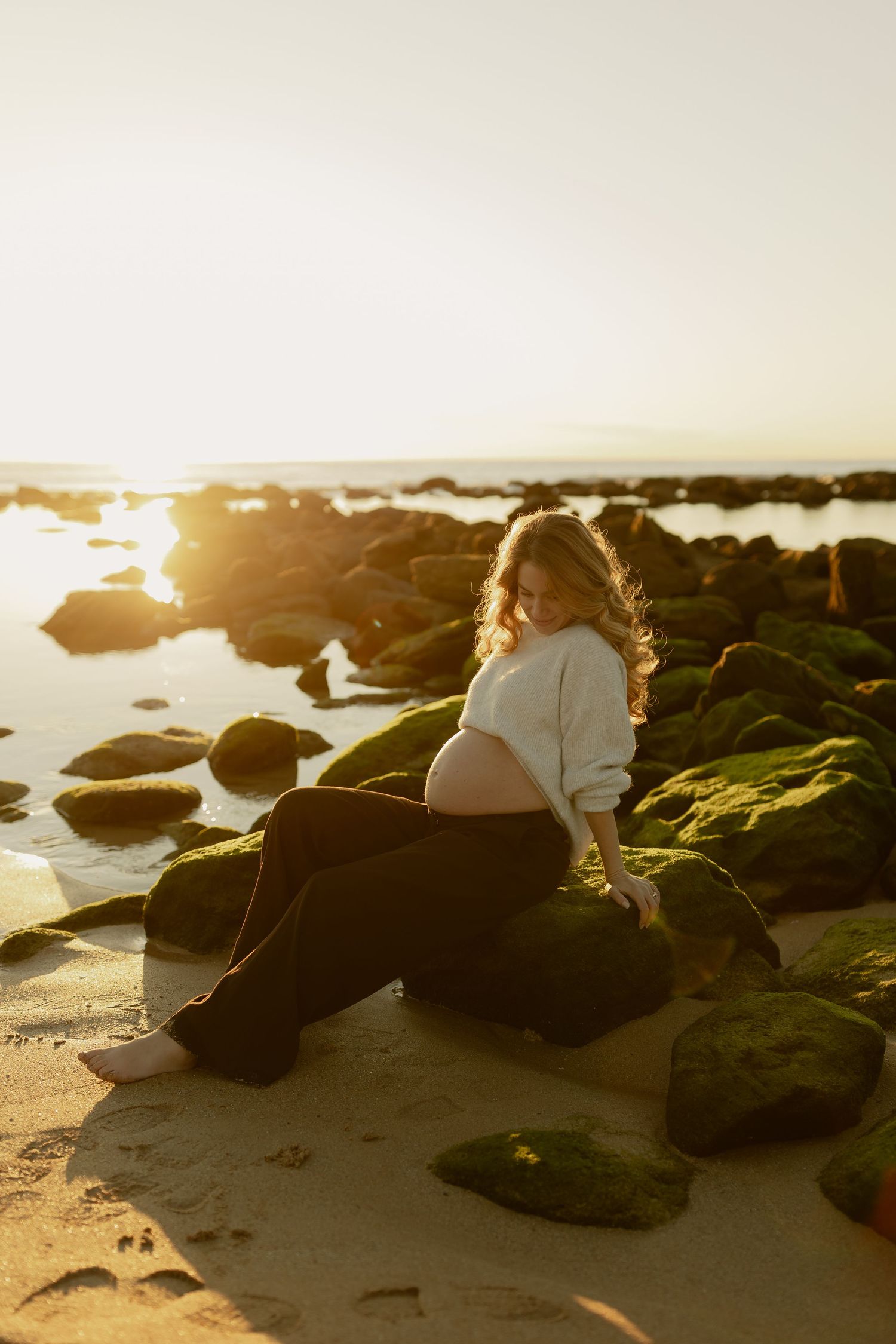 Pregnant woman sitting on moss-covered rocks at beach during golden sunset, cradling her baby bump in a white sweater.