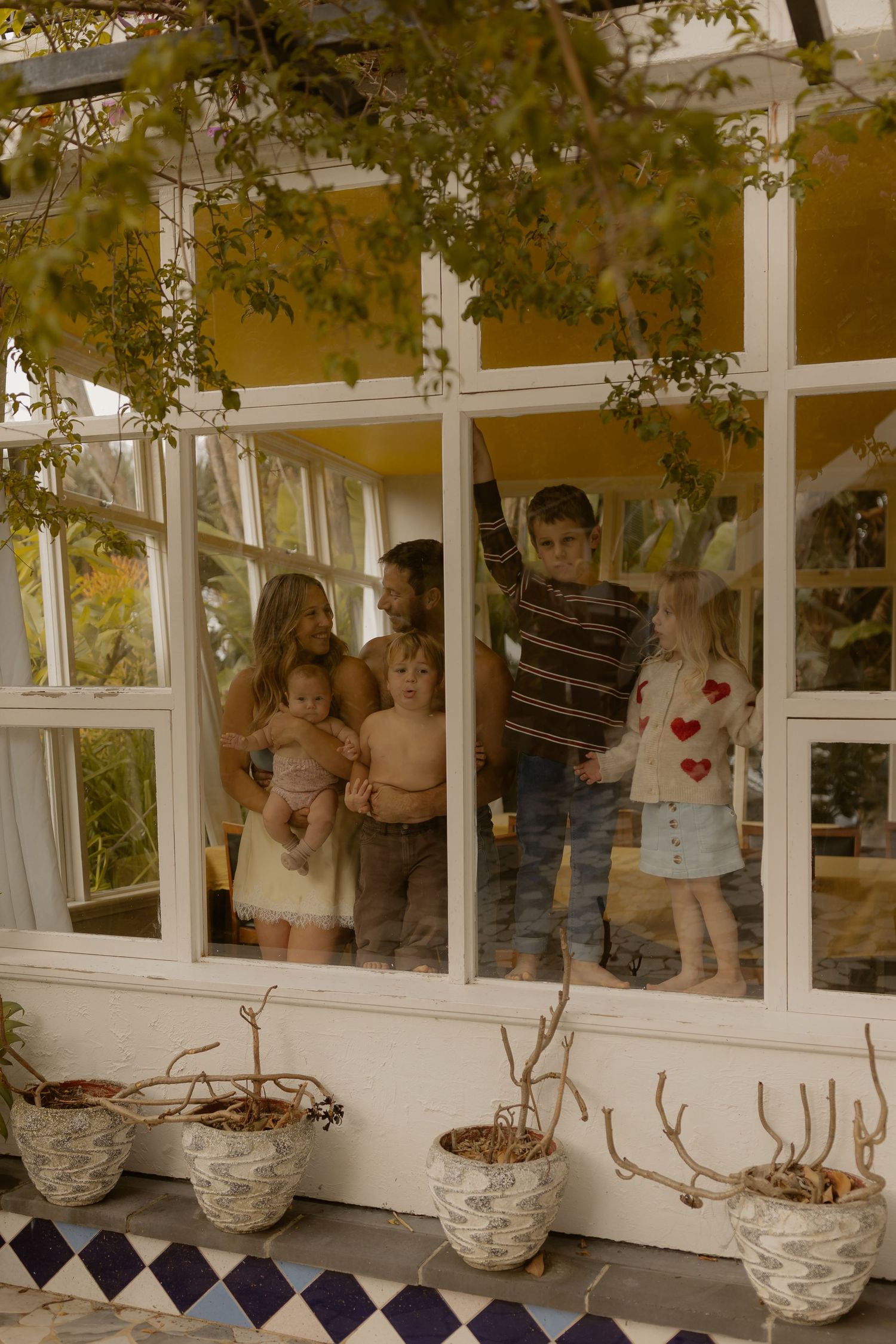 Family of five children and adults standing together inside a sunlit greenhouse with white window frames and potted plants.