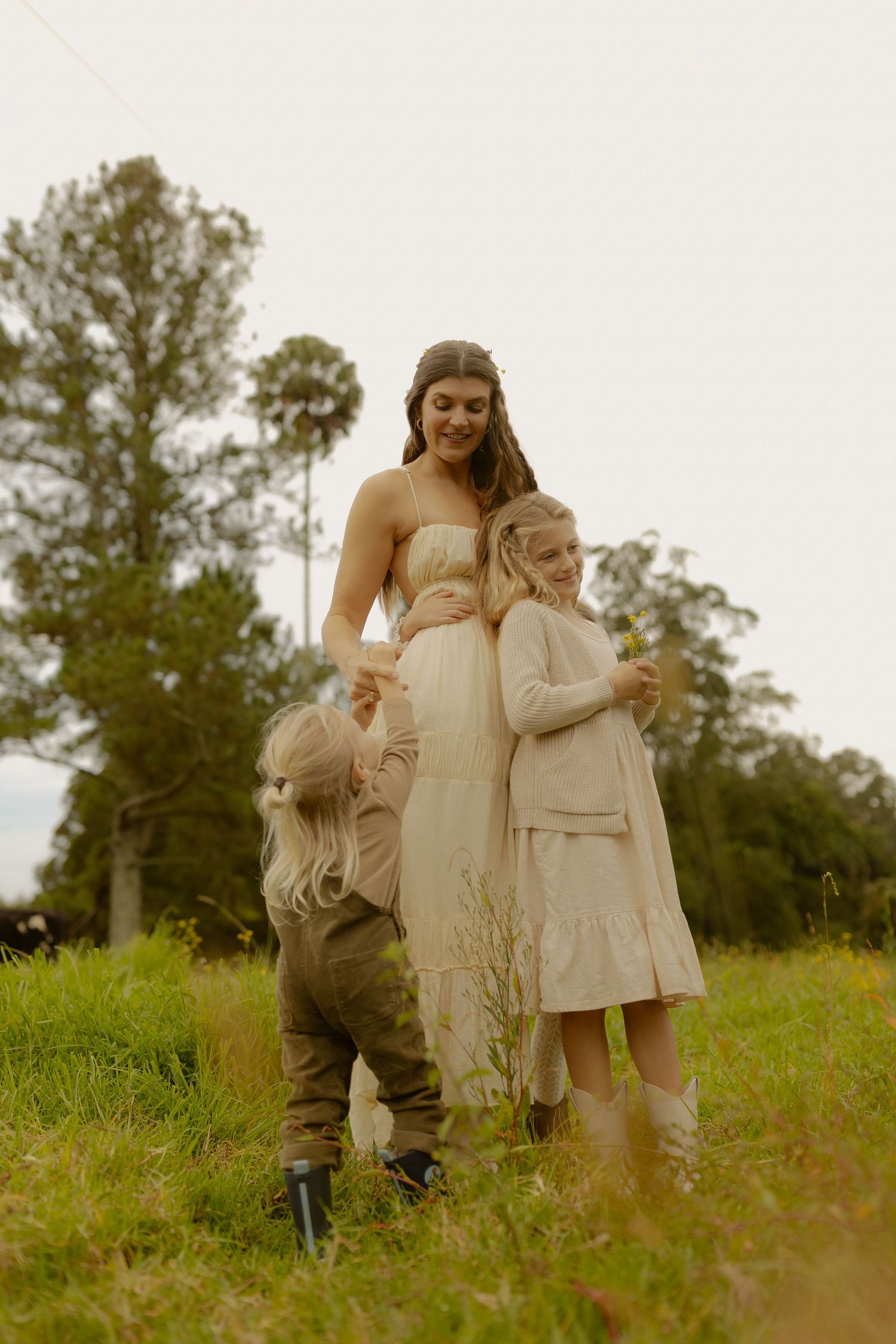 Pregnant woman in cream dress smiling at two young children hugging her bump in a green field.