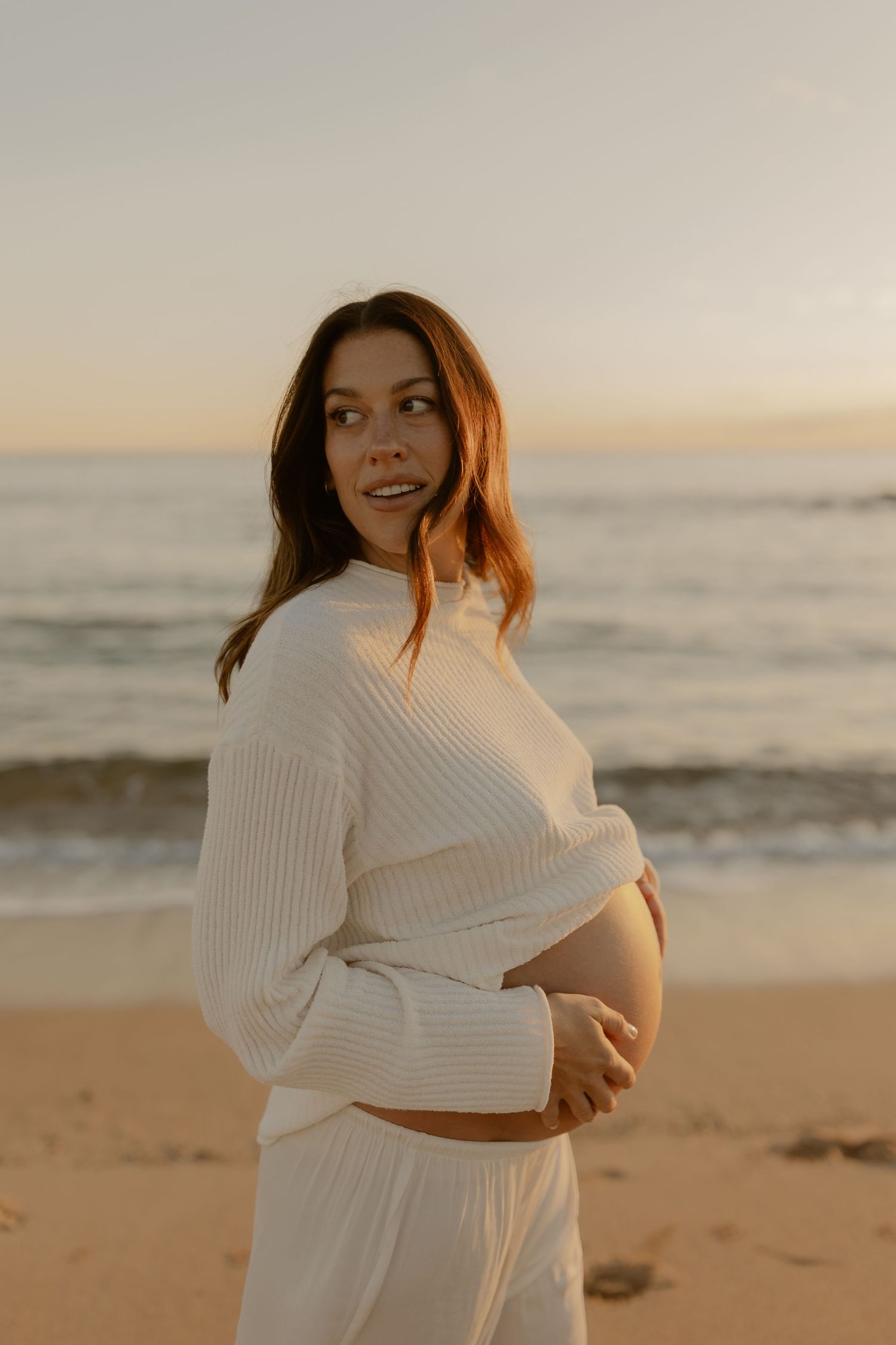 Pregnant woman in white ribbed sweater cradling baby bump on sandy beach at golden sunset.