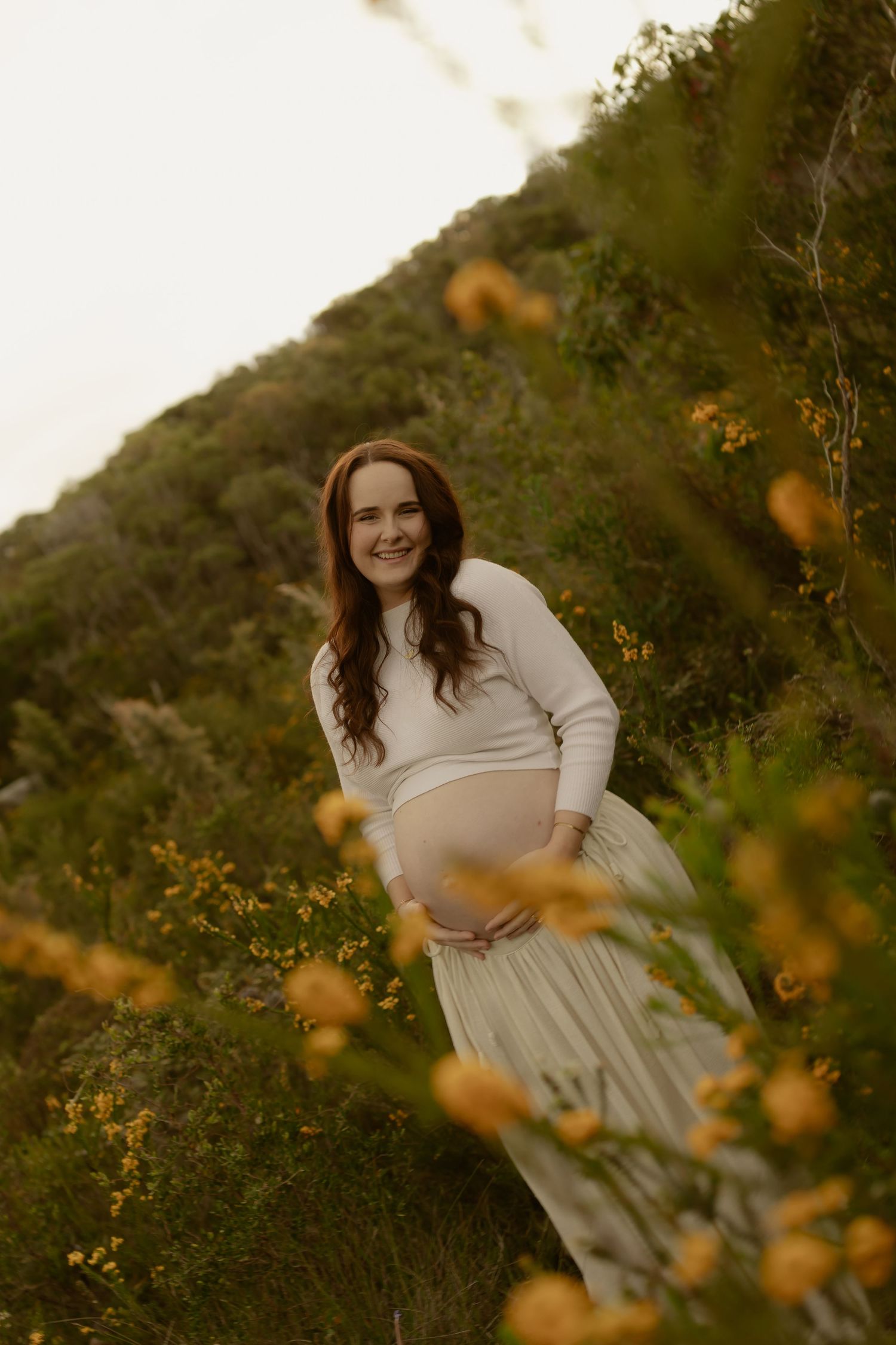 Smiling pregnant woman in white top and skirt cradling baby bump outdoors among yellow wildflowers and green hills.