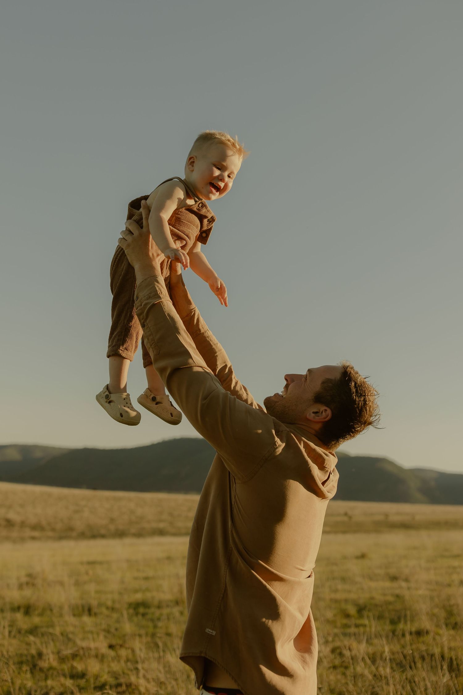 A father joyfully lifts his laughing toddler high in the air in a golden field at sunset.