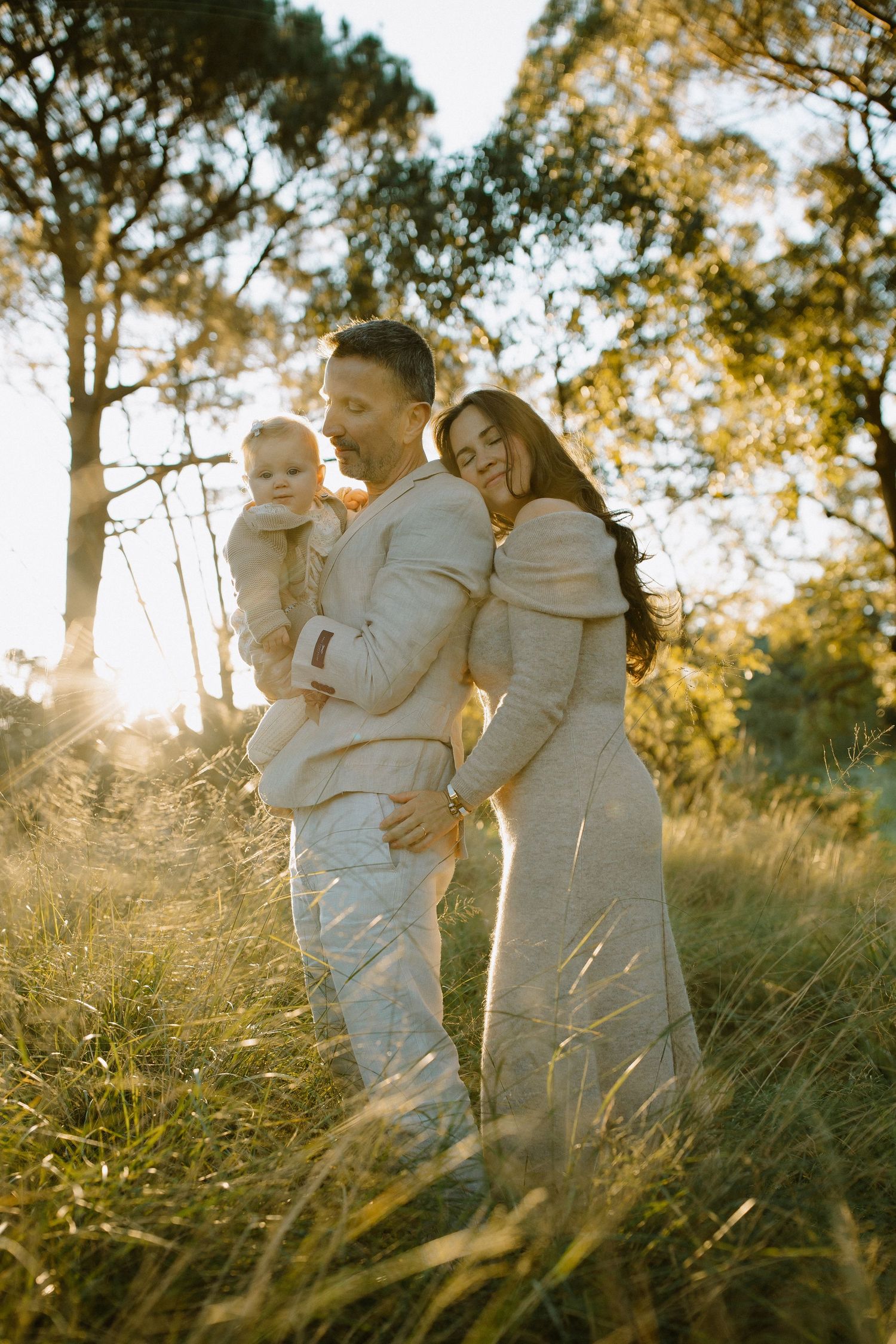 Family in matching light outfits poses together in sunlit field during sunset.