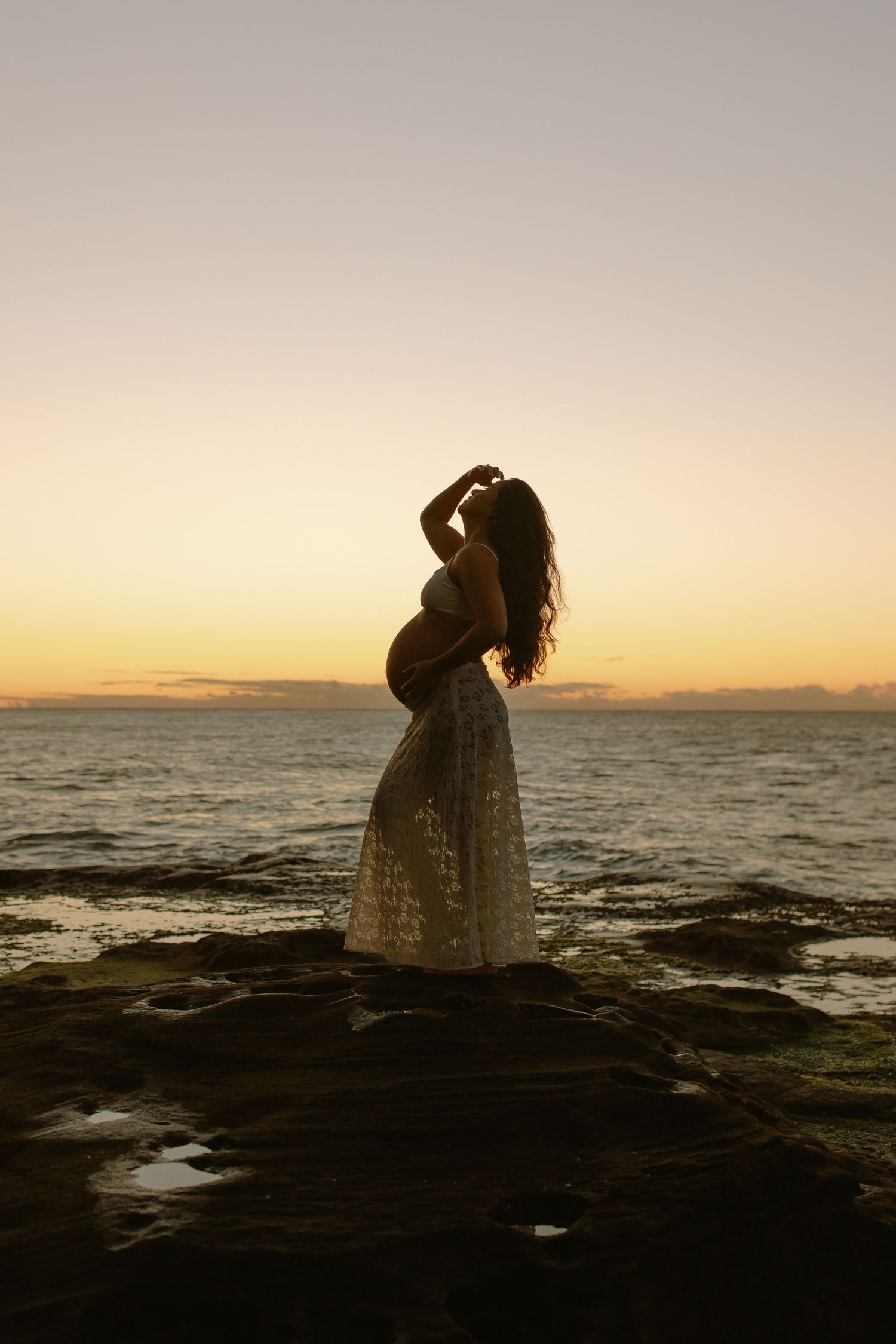 Silhouette of a pregnant woman in a lace dress standing on rocky shore during golden sunset over ocean waters.
