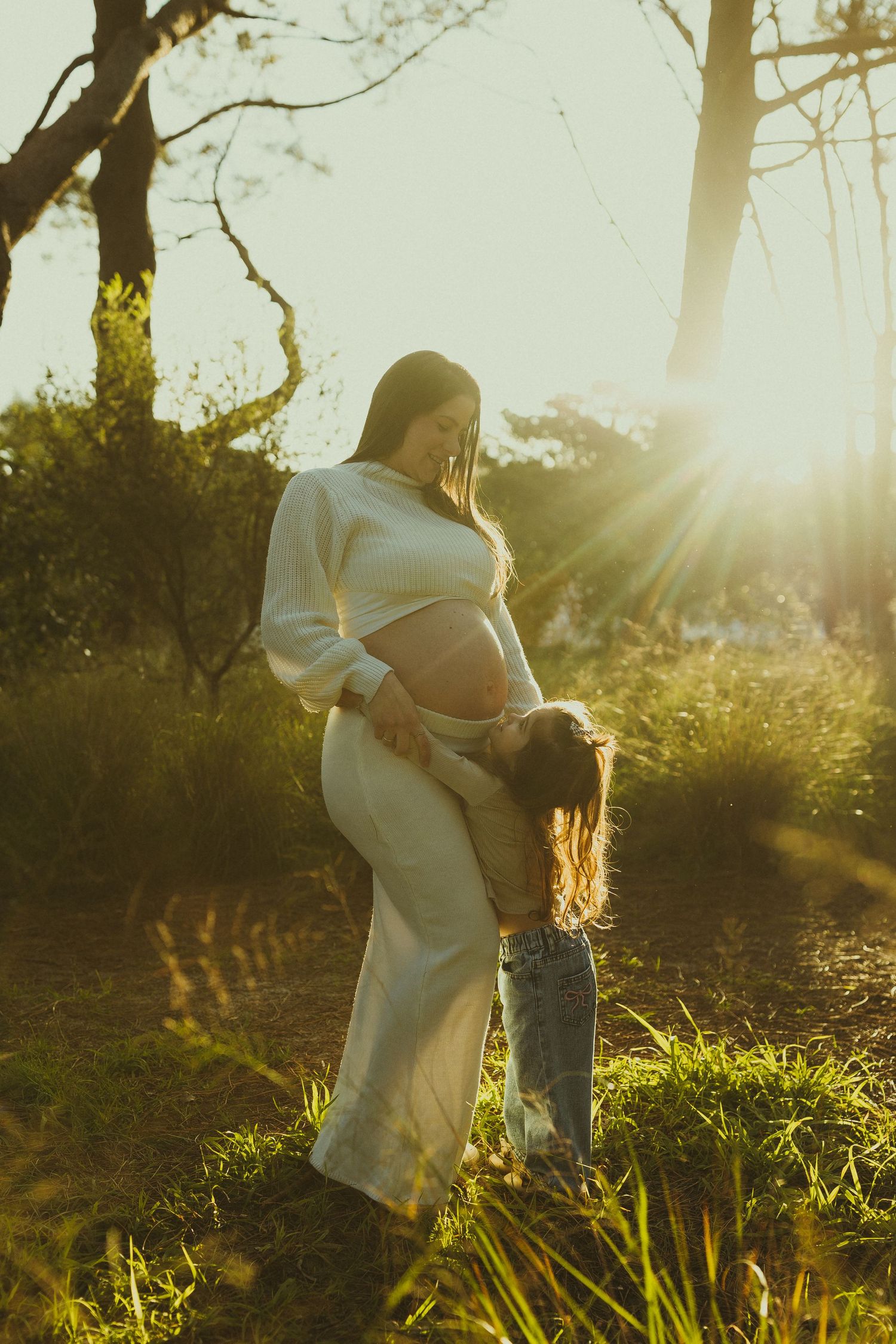 Beautiful sunset maternity photoshoot in a natural outdoor setting with tall grass and streaming sunlight.