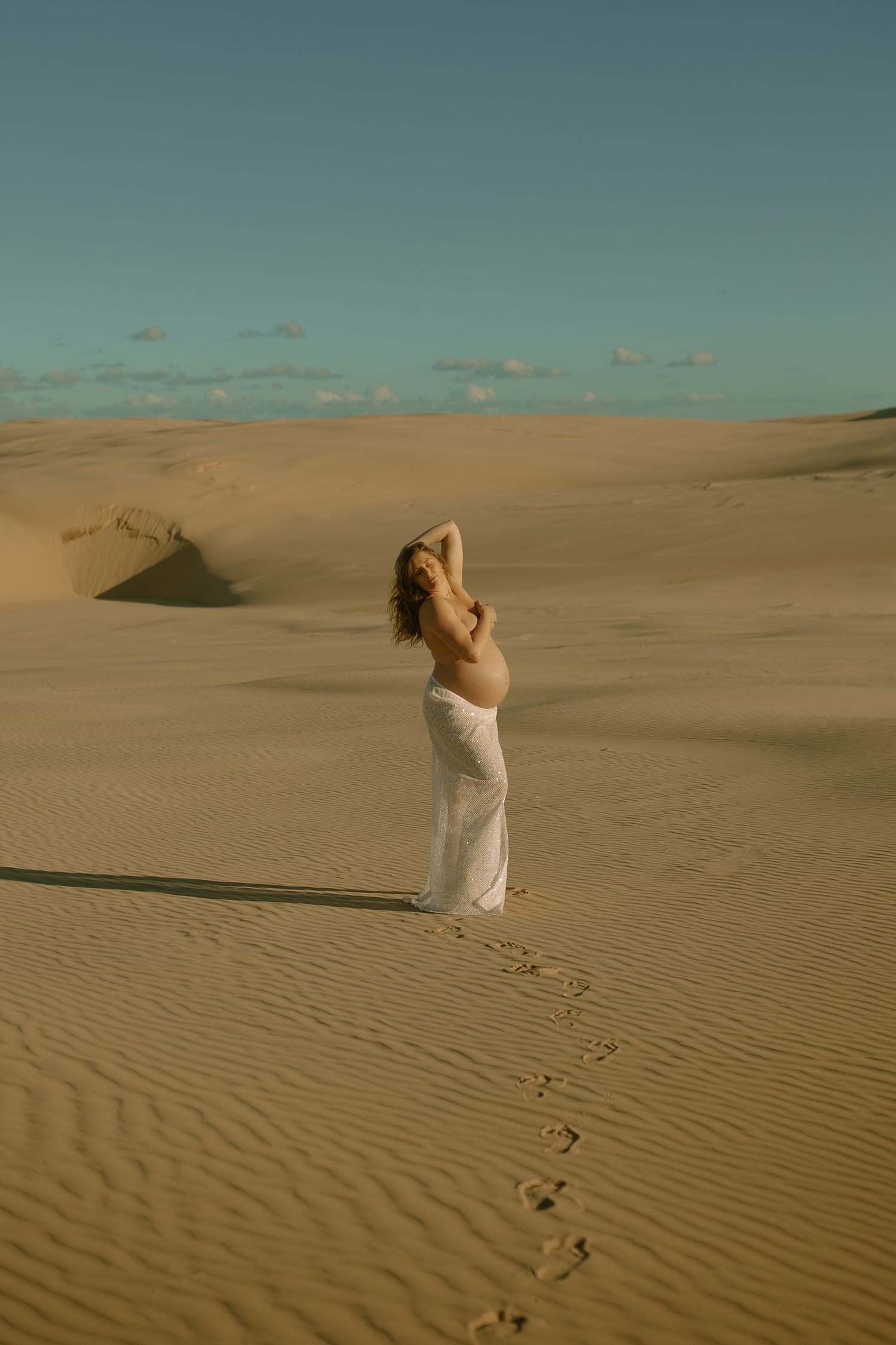 Footprints trail behind a white-dressed figure standing among vast desert sand dunes under a turquoise sky.