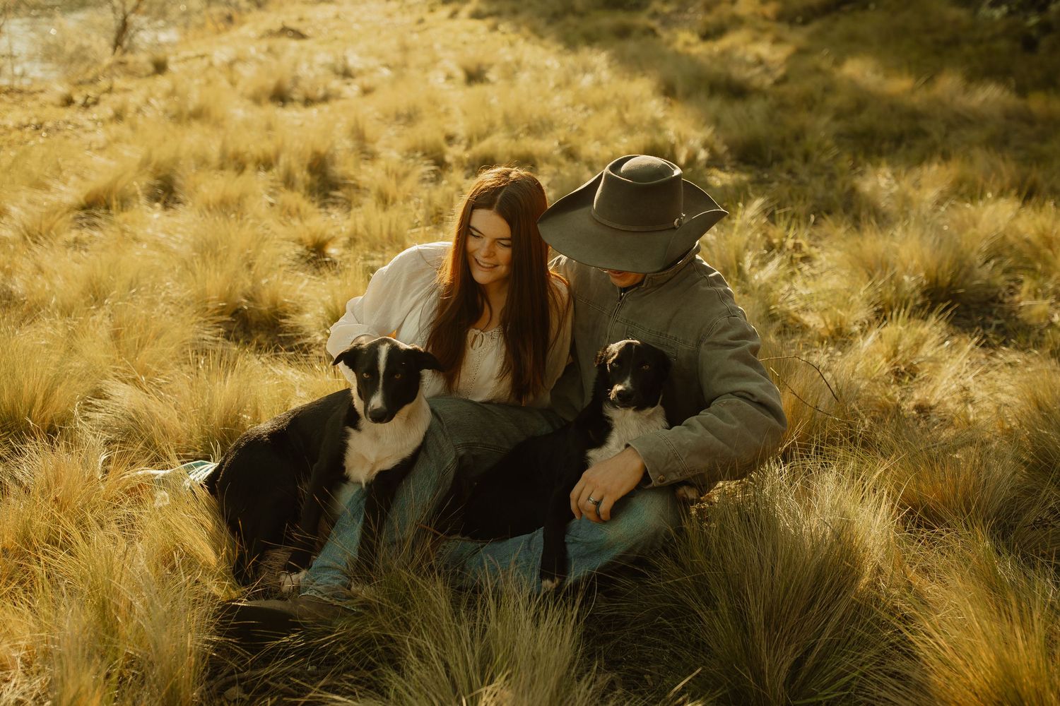 A couple sits together in golden grass during sunset with their black dog beside them.