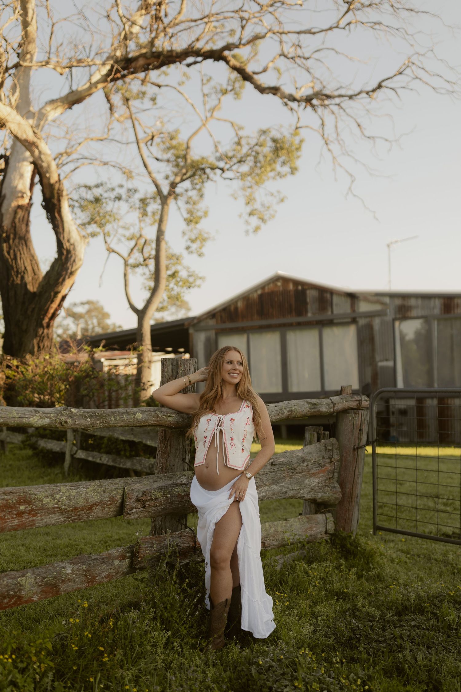 A smiling woman in a white dress and brown boots leans on a rustic wooden fence at golden hour on a farm.
