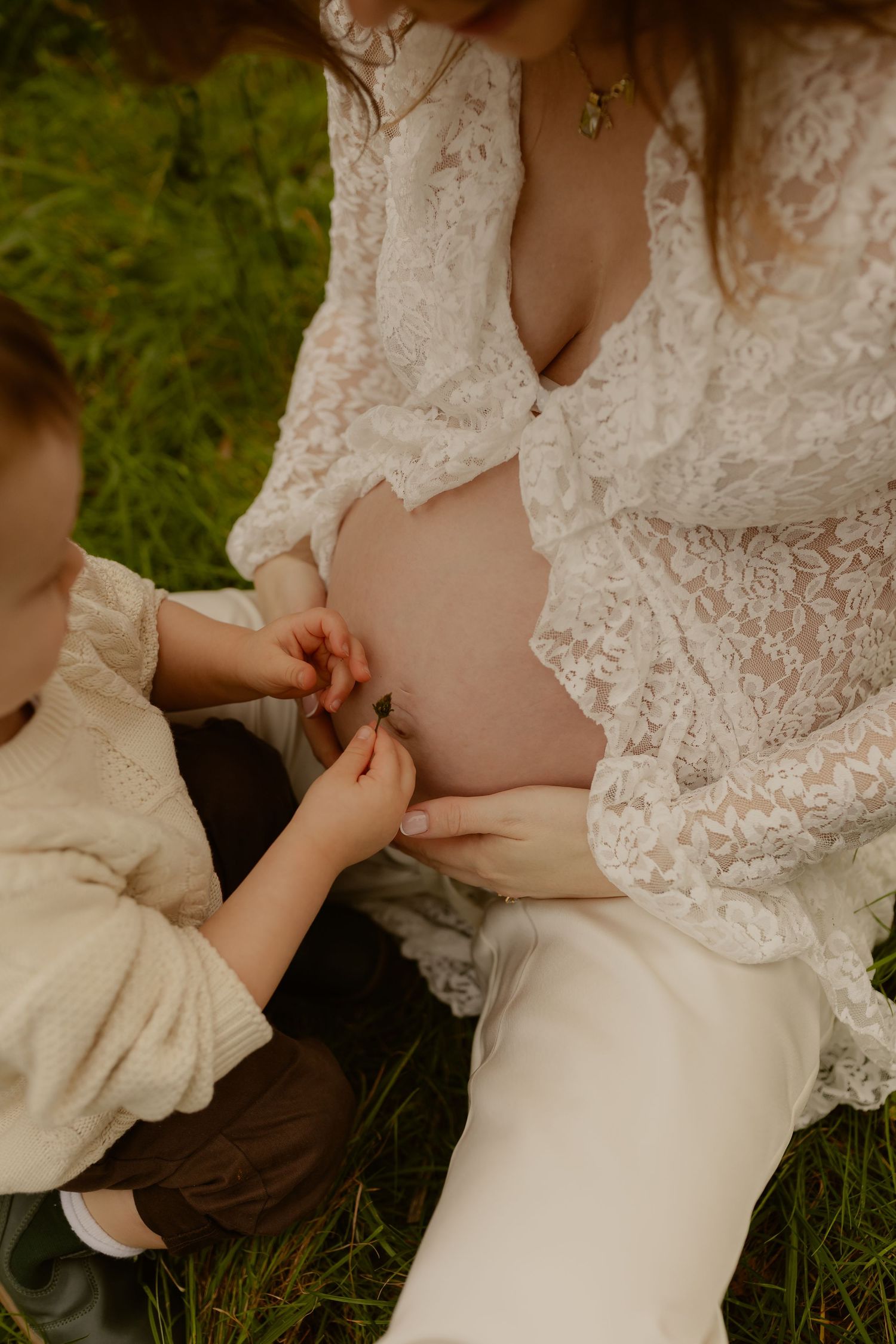 A young child touches a pregnant woman's bare belly outdoors during a maternity photoshoot.