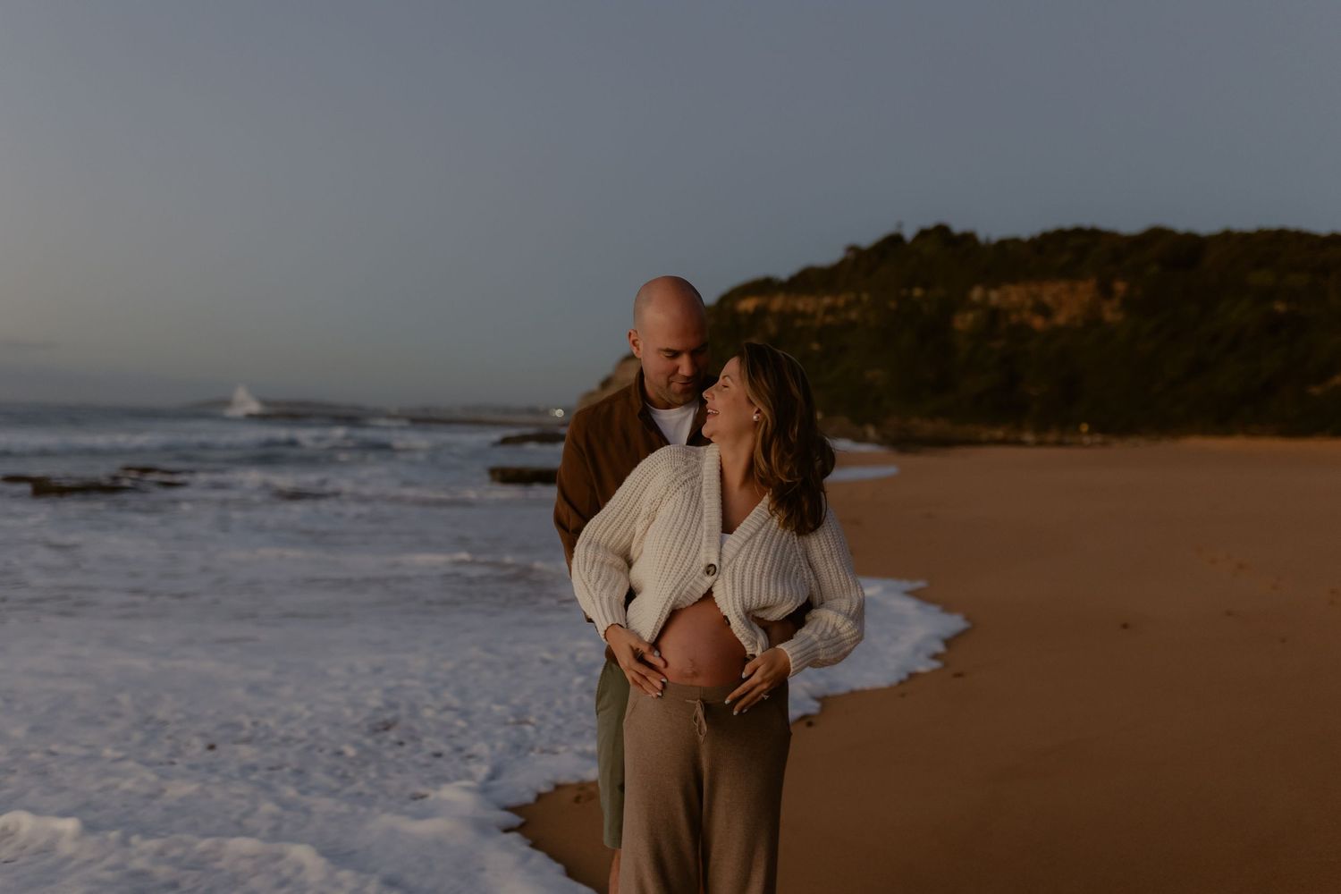 Couple embracing on sandy beach at dusk, woman cradling baby bump as waves wash ashore near rocky cliffs.