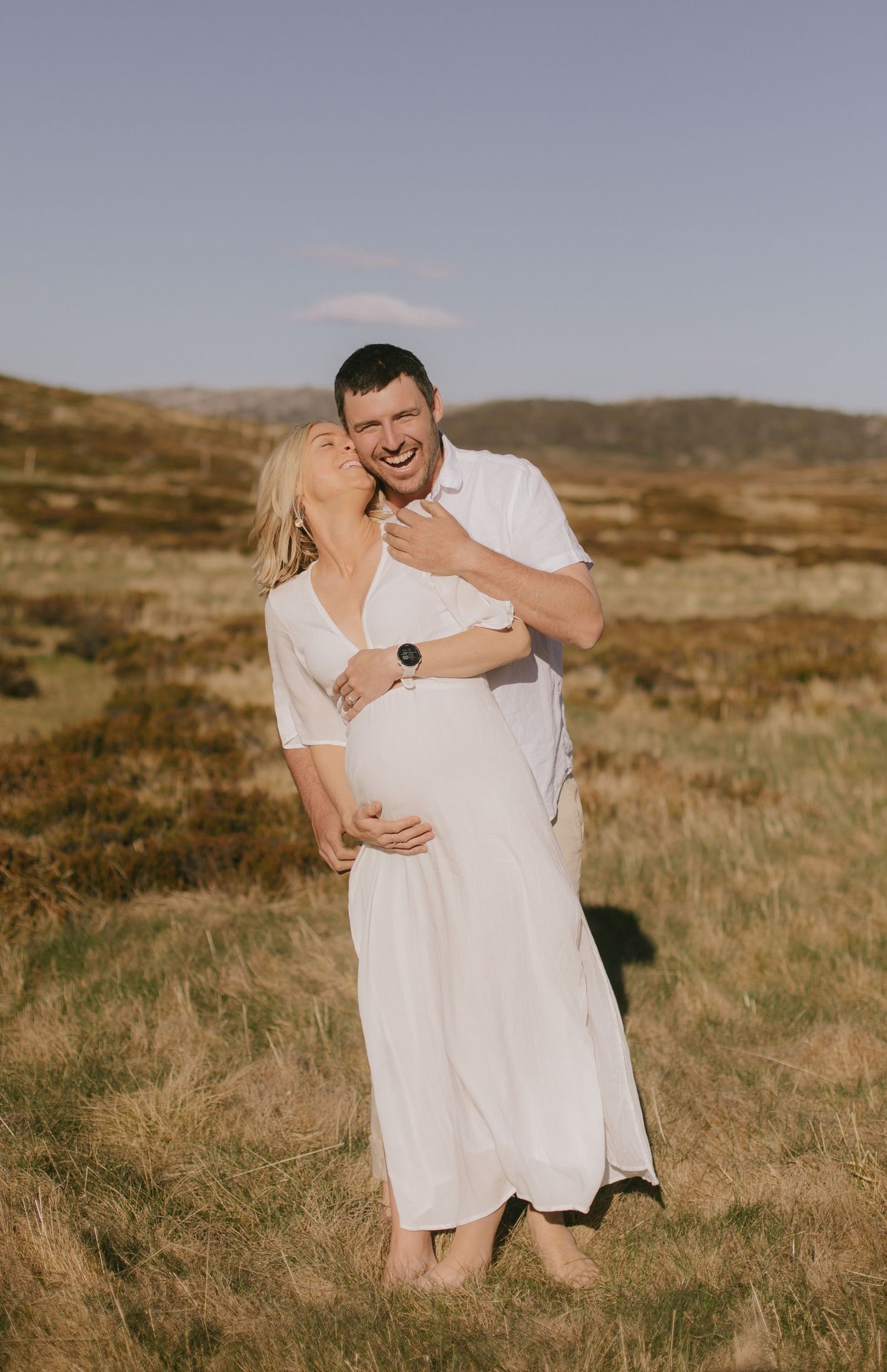 A couple in white outfits embrace and laugh together in a golden field with rolling hills in the background.