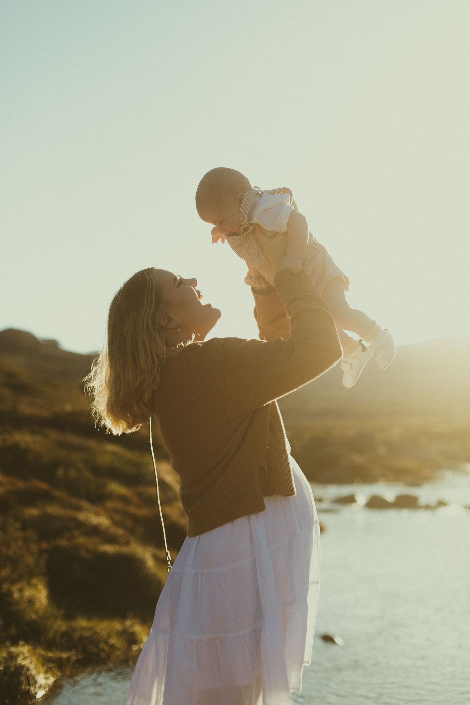 Silhouette of parent lifting child against golden sunset sky with ocean coastline and rocks in background.