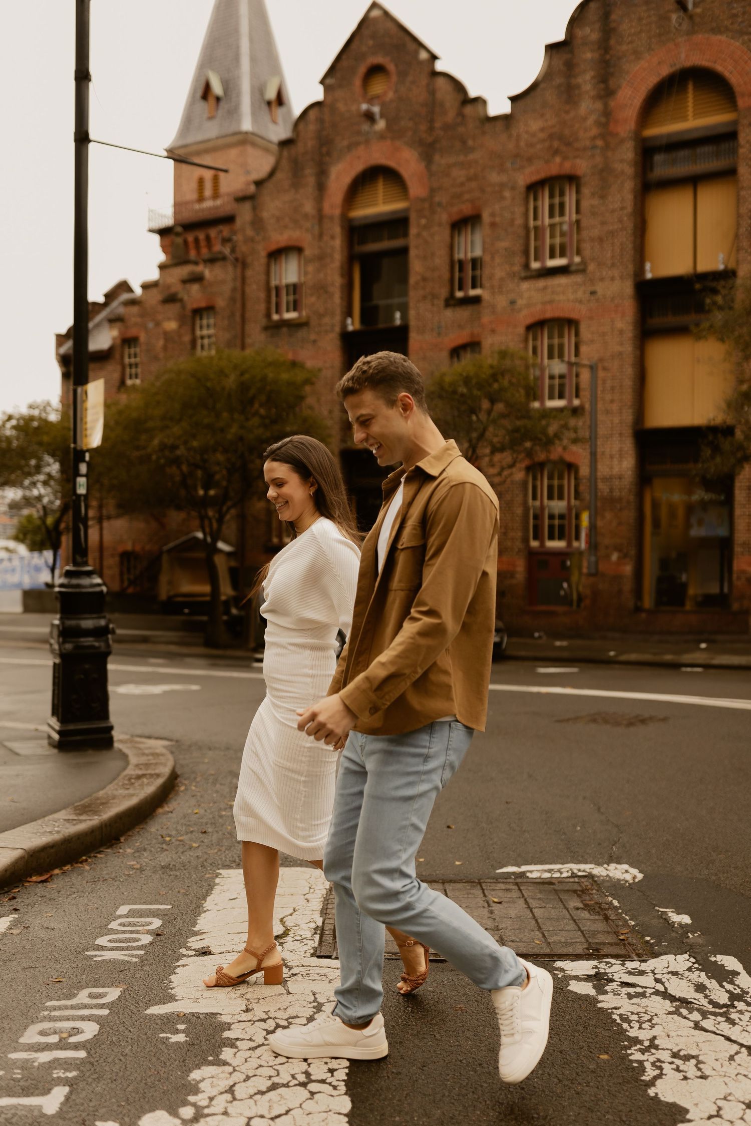 A couple walks along a cobblestone street with historic brick buildings and church spires in the background.