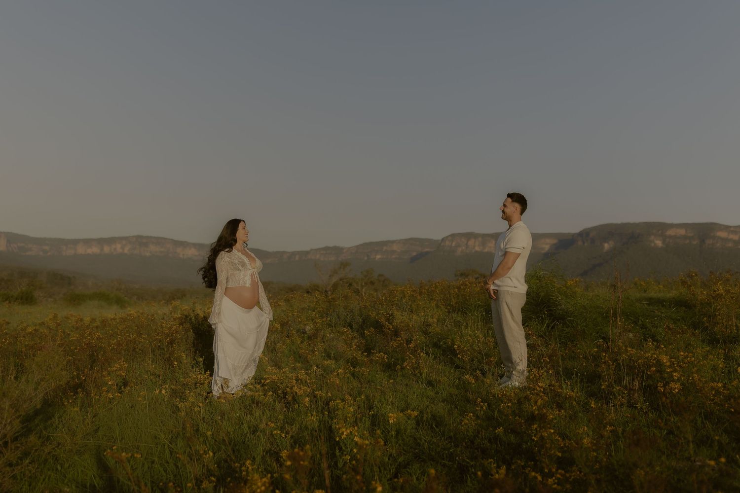 People in white dresses walk through a golden field at sunset with mountains in the background.