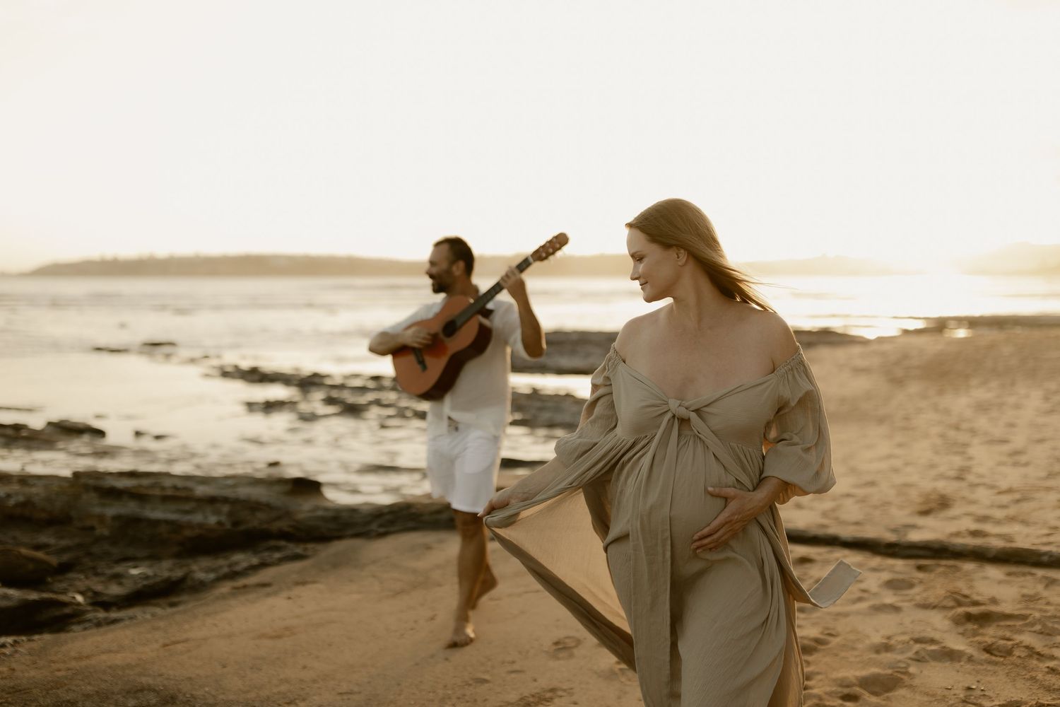 Two people in light colored clothing dance playfully on a beach during golden hour.