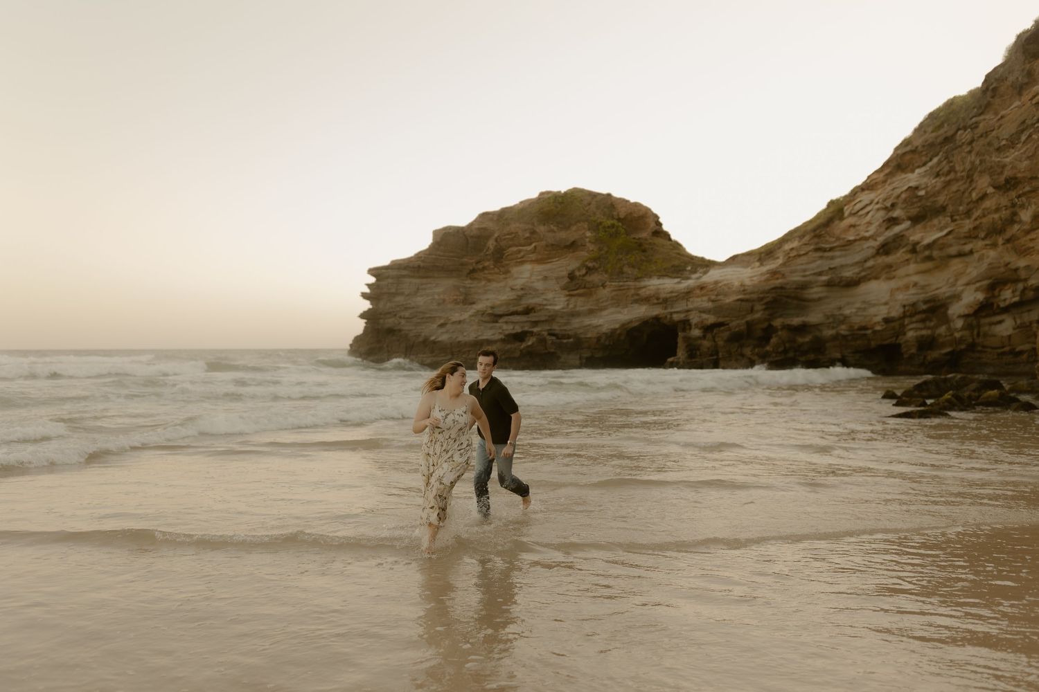 Couple exploring a dramatic rocky coastline with waves crashing against cliffs at sunset.