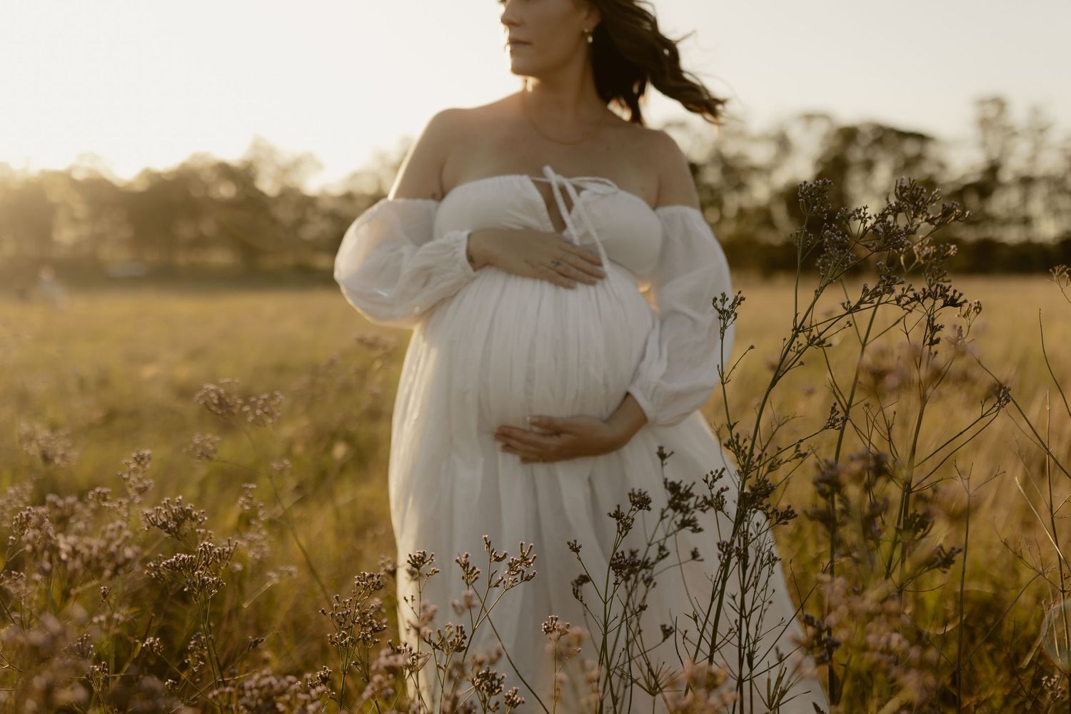 A dreamy maternity photo of a flowing white dress in a golden sunlit field at sunset.