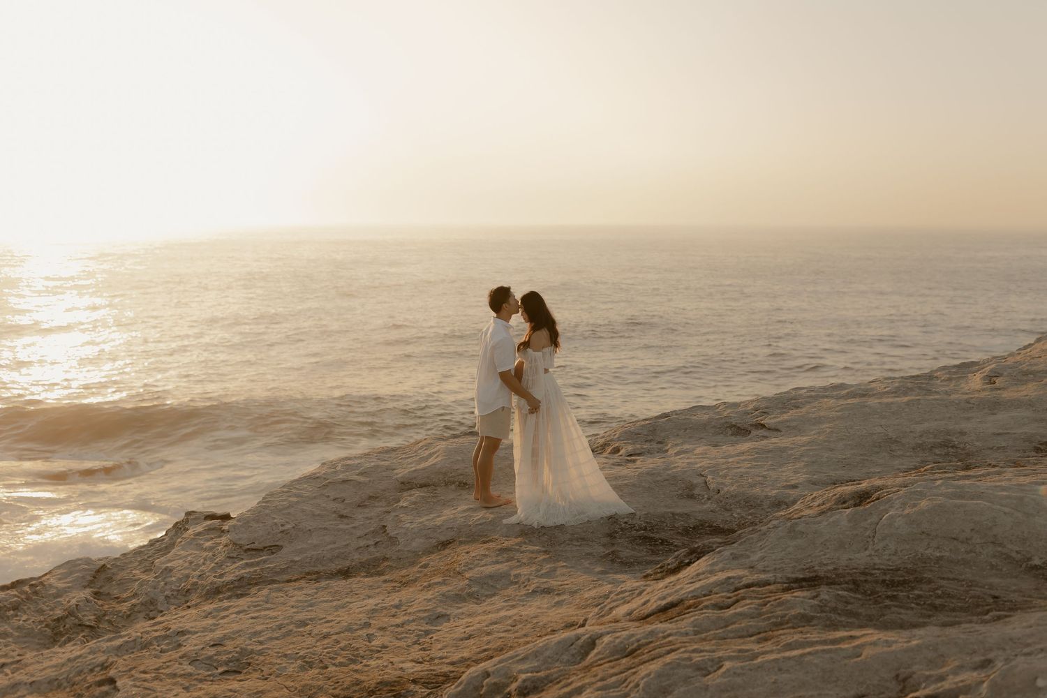 Couple in white wedding attire embracing on rocky coastal cliffs at sunset with ocean waves crashing below.
