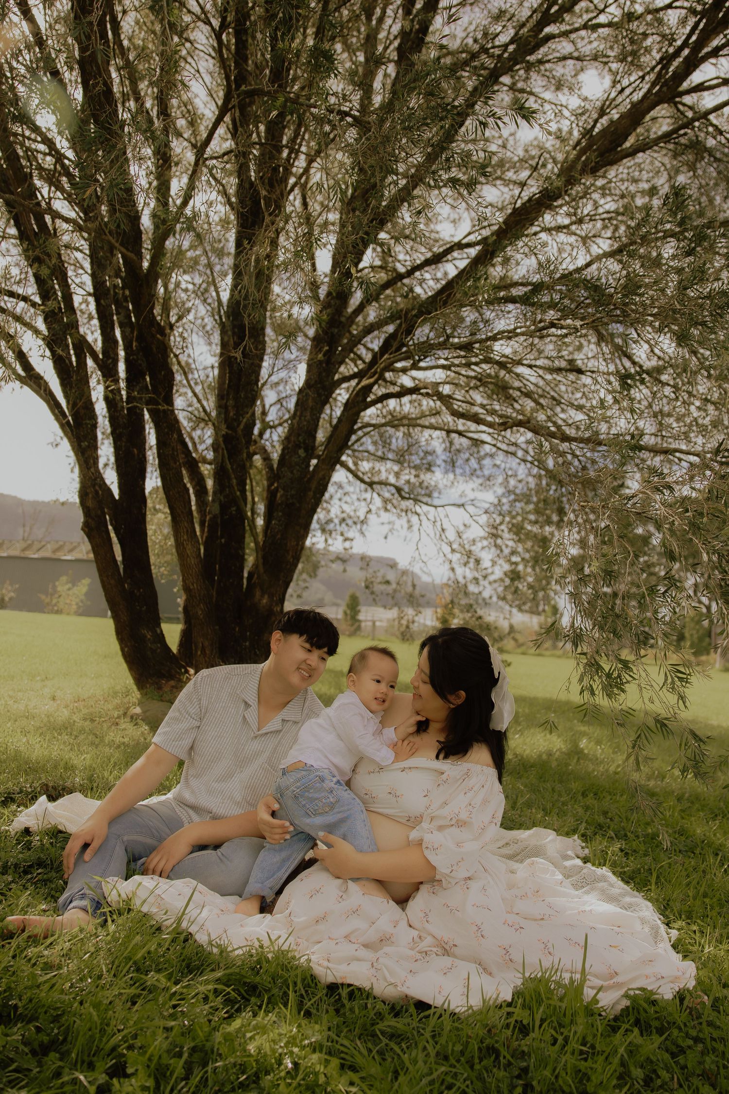 Family sitting on a blanket under a willow tree in a green park, parents smiling at their young child.