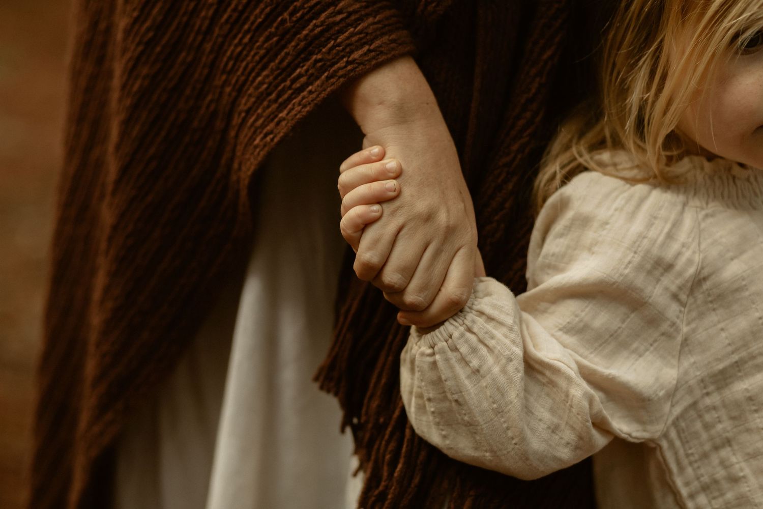 Close-up detail of a small hand grasping a brown knit fabric against a cream-colored linen background.