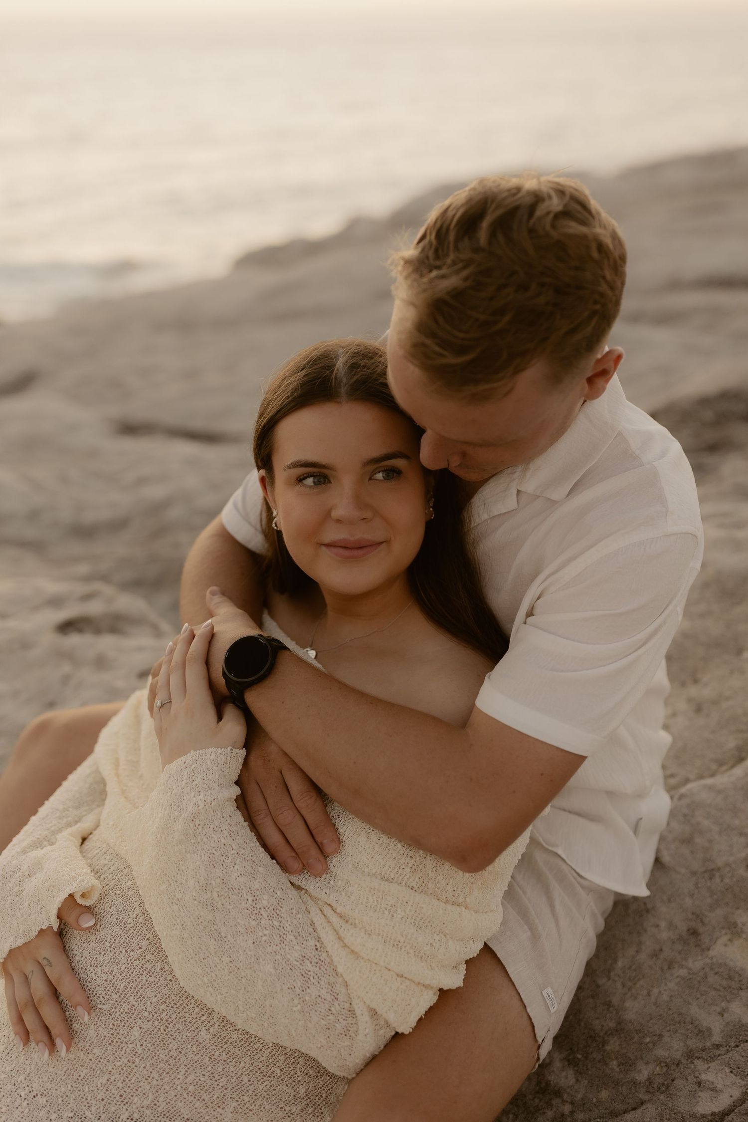 A romantic couple embraces on the beach at sunset wearing white and cream colored outfits.