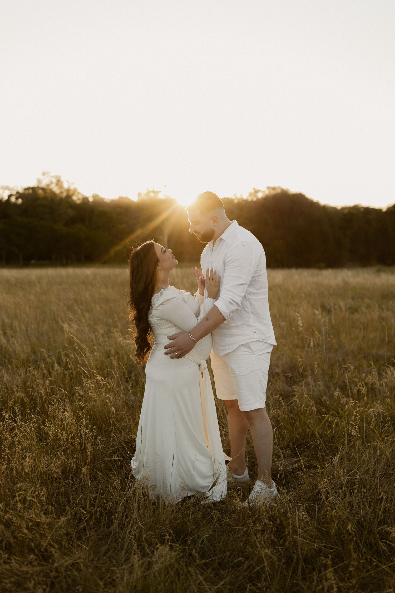 A romantic sunset maternity photo shows a couple embracing in a golden field wearing white outfits.