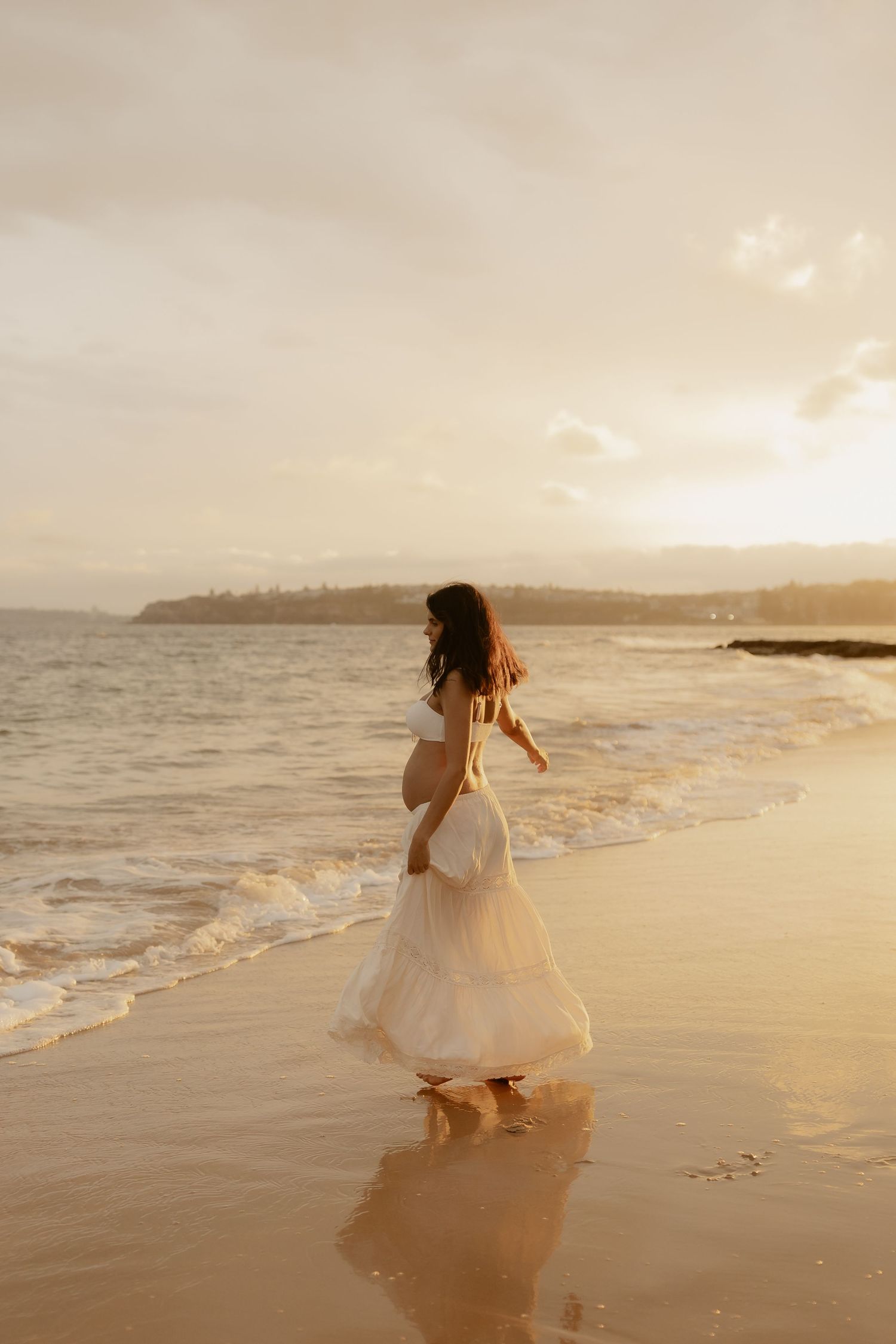 Person in a flowing white dress walks along a sunset beach with waves rolling in.