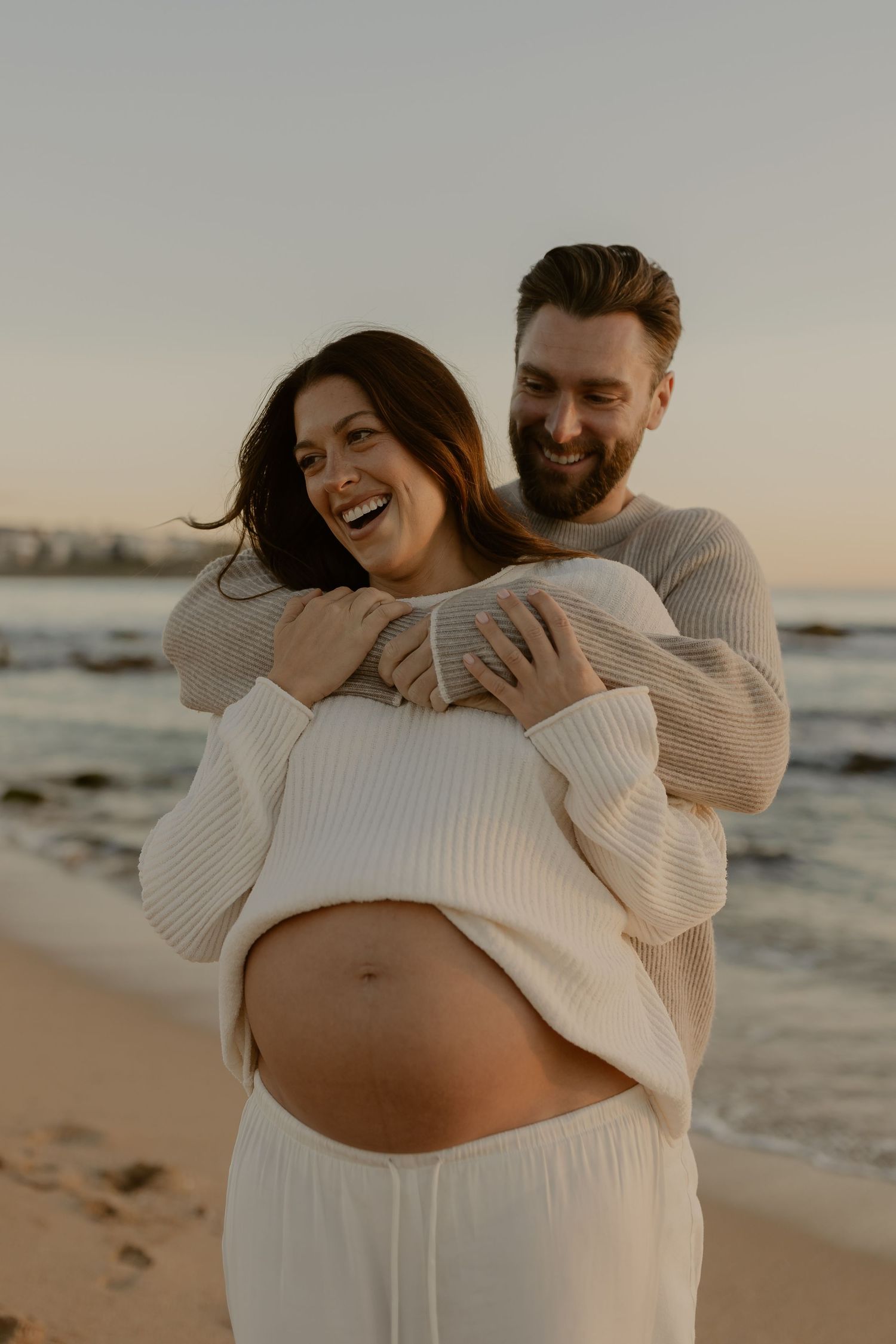 Joyful couple in matching sweaters share intimate moment during sunset beach maternity session.