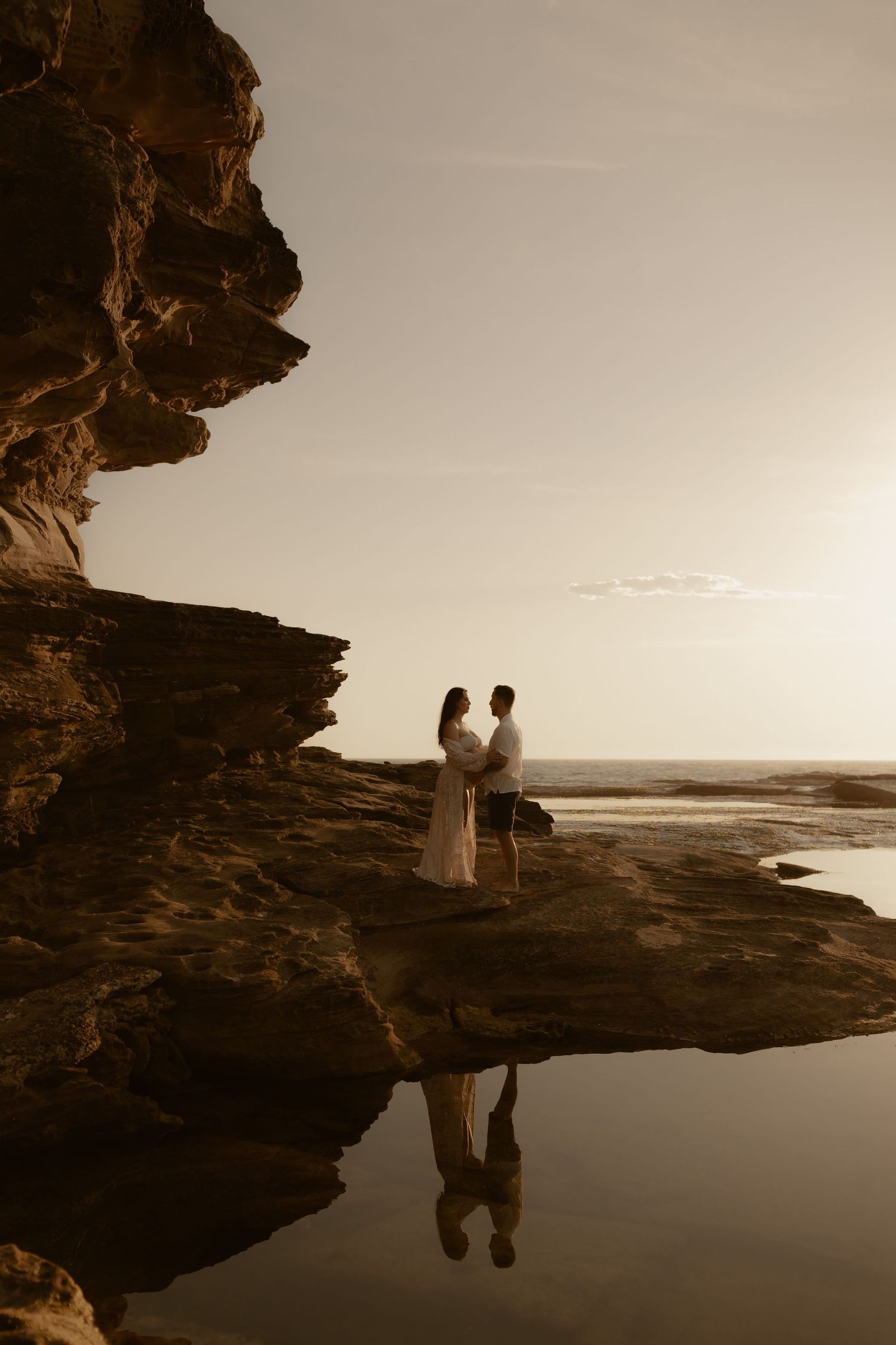 A couple stands silhouetted against the sunset on a rocky coastal outcrop with their reflection in a tidal pool.