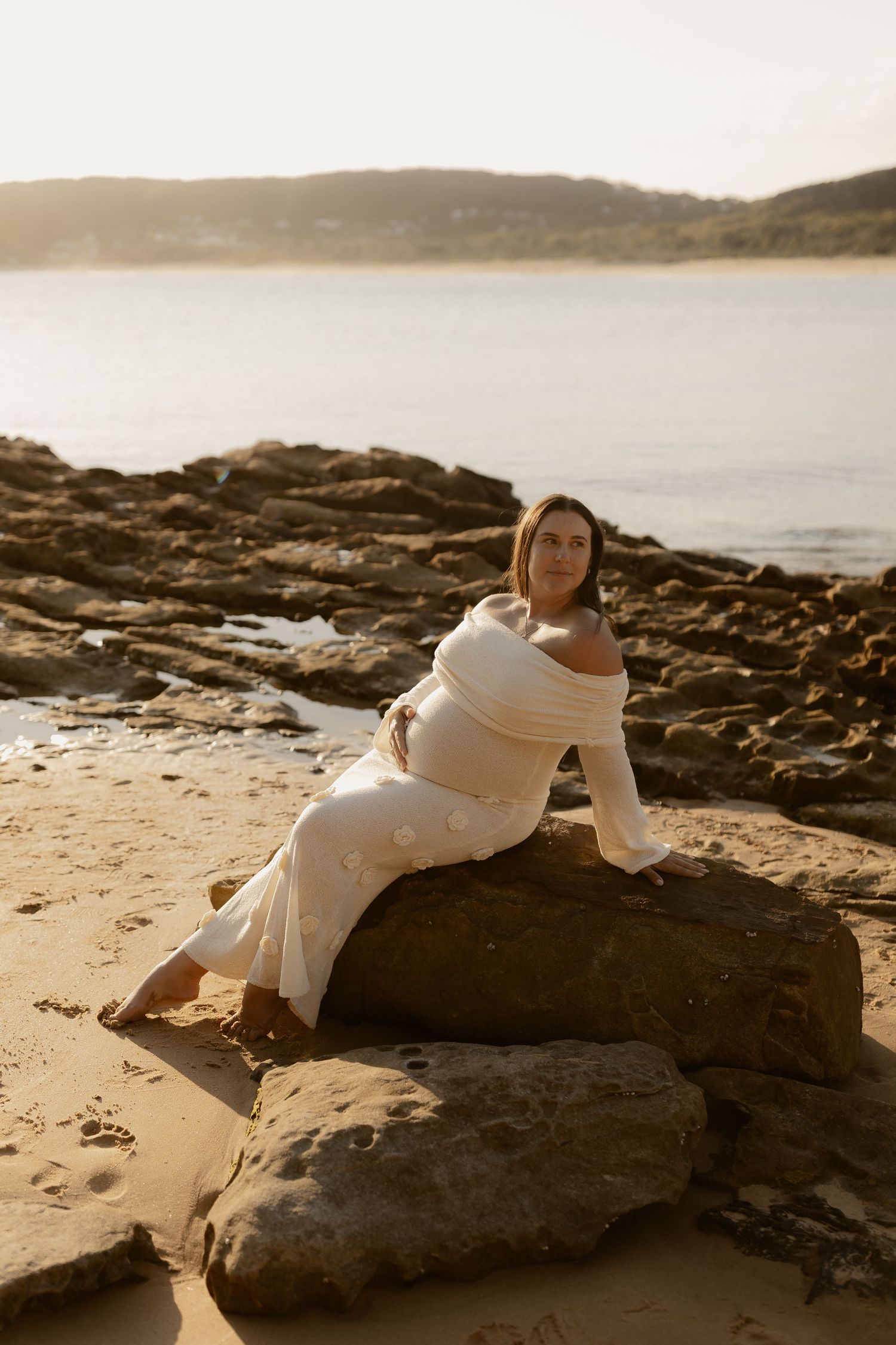 Person in white dress lying on coastal rocks during a sunset photoshoot by the ocean.