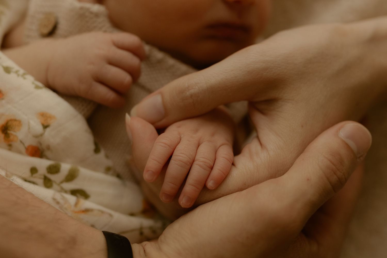 A tiny baby hand gently grasps an adult's finger against a soft floral fabric background.