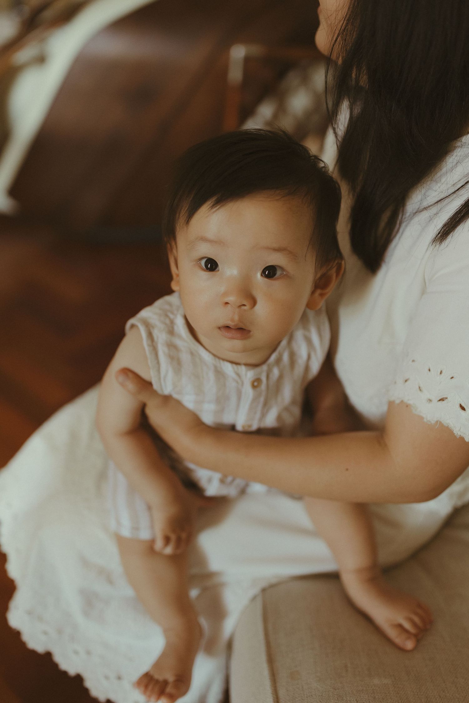 A baby in white clothing sits on someone's lap in a warmly lit room.