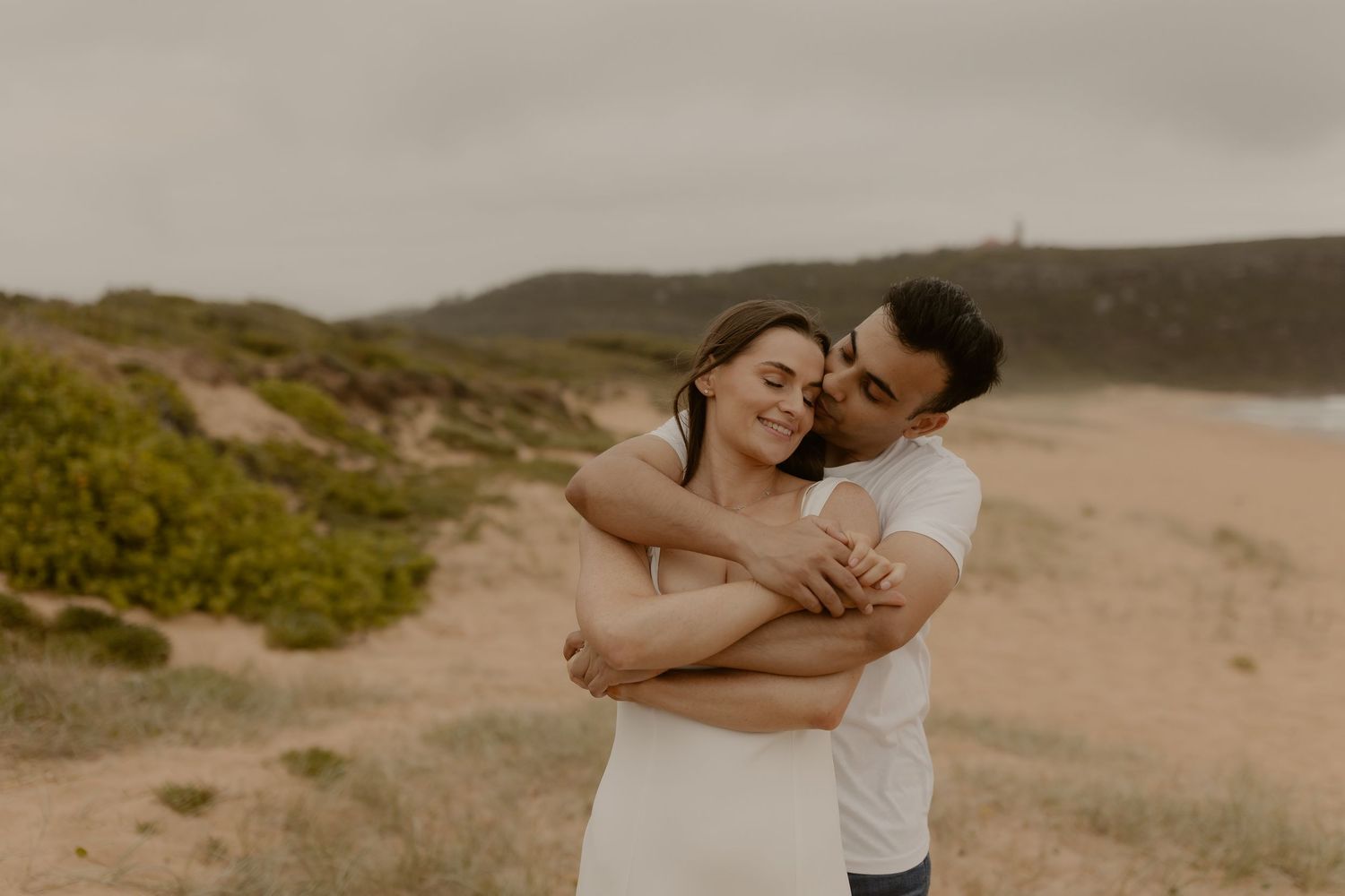 Couple embracing on coastal cliffs with dramatic ocean landscape in warm muted tones.