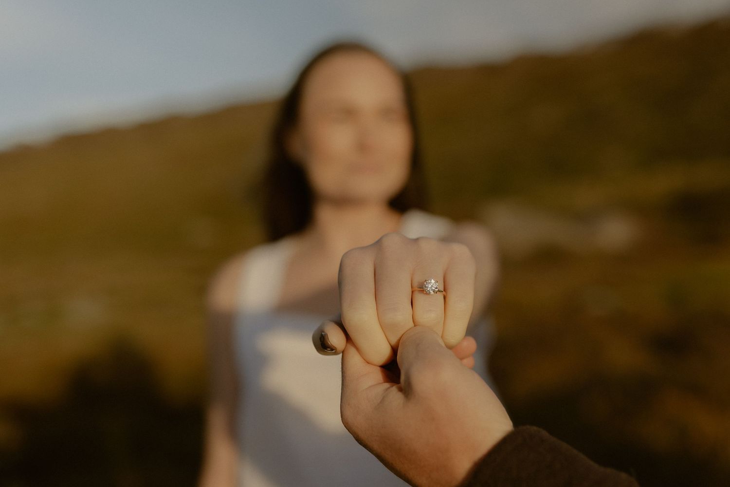 Person holding up engagement ring in golden sunlight against blurred natural outdoor background.