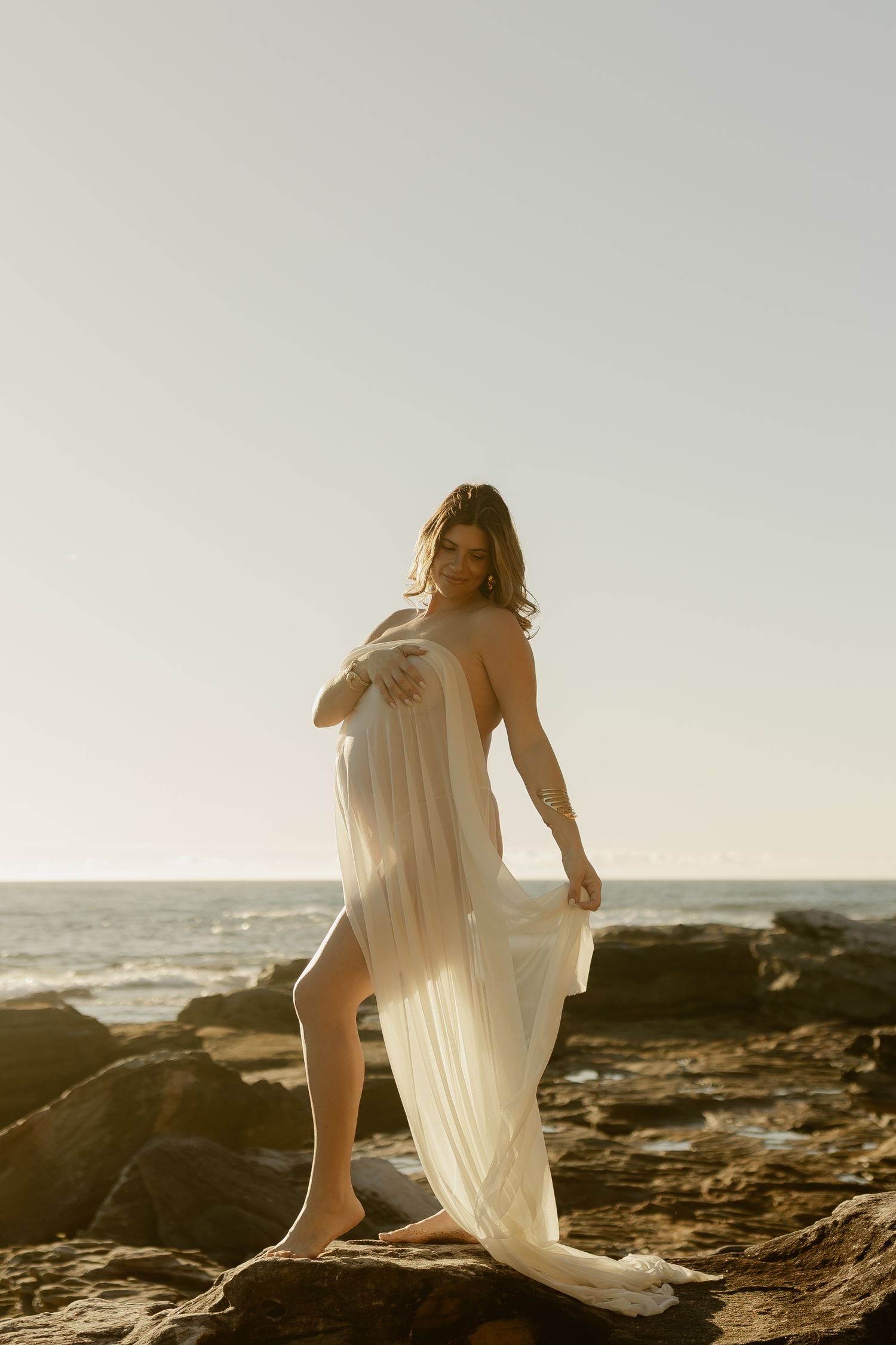 Woman in flowing white dress standing on coastal rocks at golden hour with ocean backdrop.