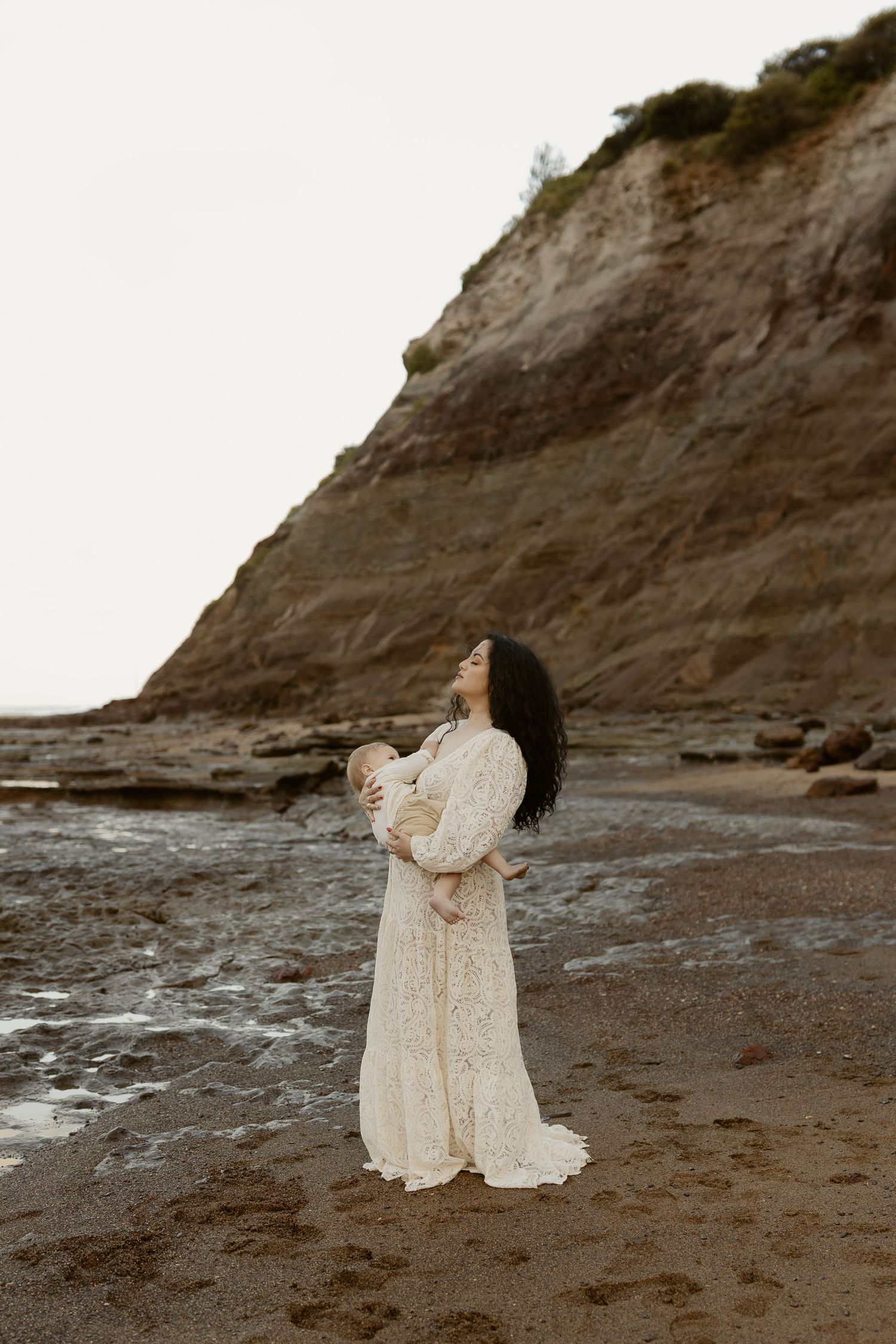 A person in a flowing white dress stands against dramatic coastal cliffs at sunset on a rocky beach.