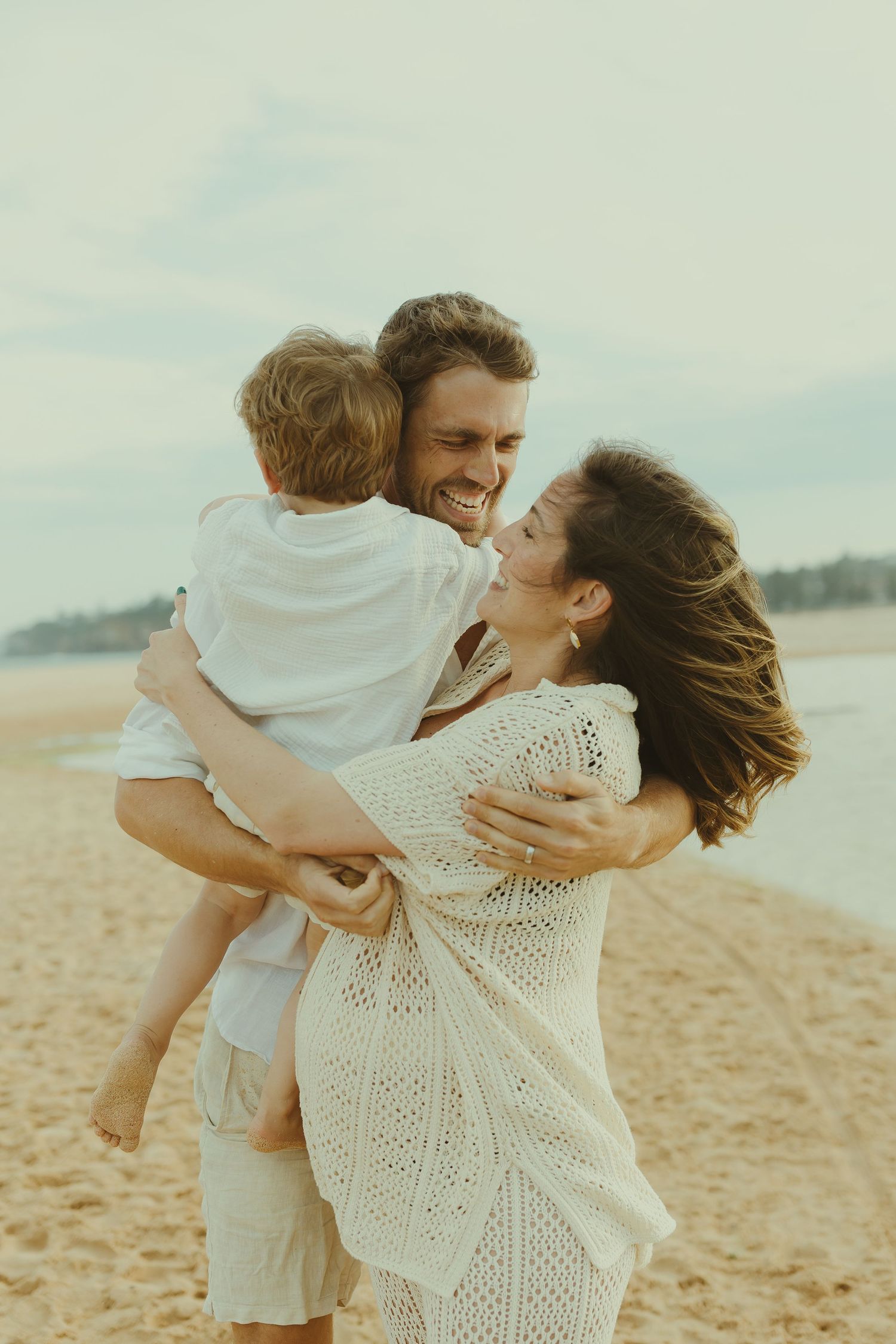A loving family shares tender moments together on a sunny beach, all dressed in white casual clothing.
