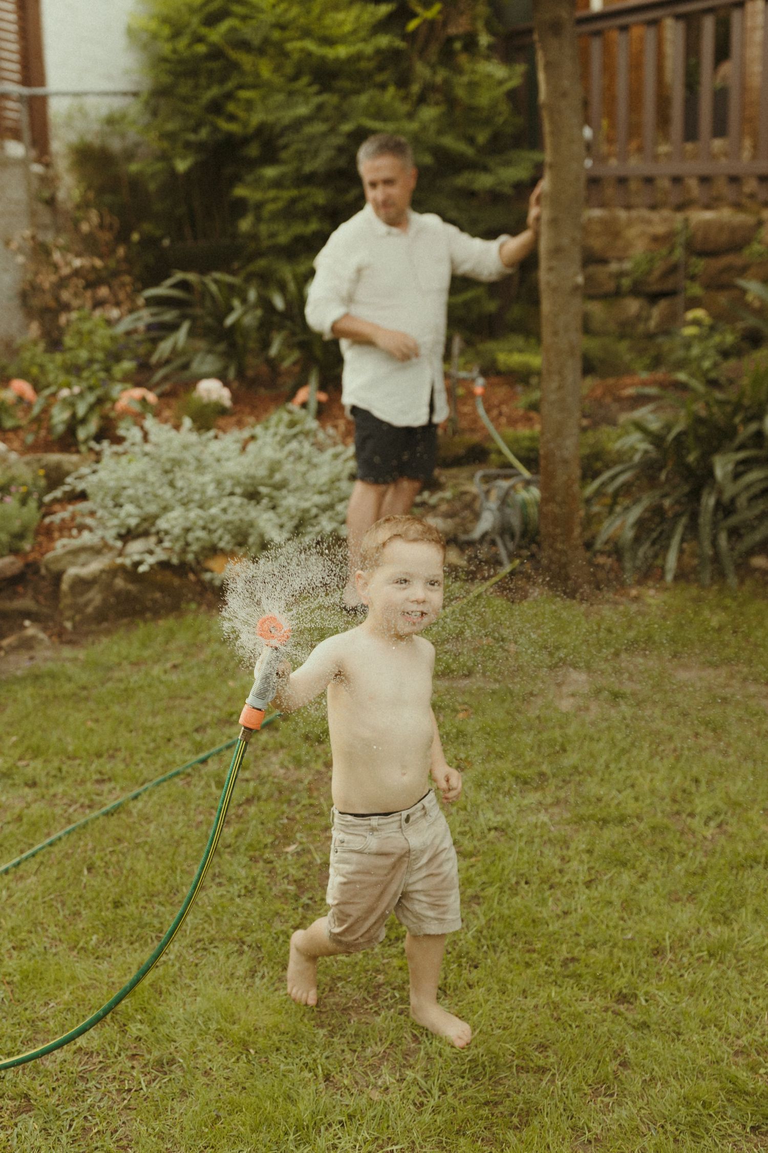 A young child plays joyfully with a garden hose on a sunny day in a backyard with trees and plants.