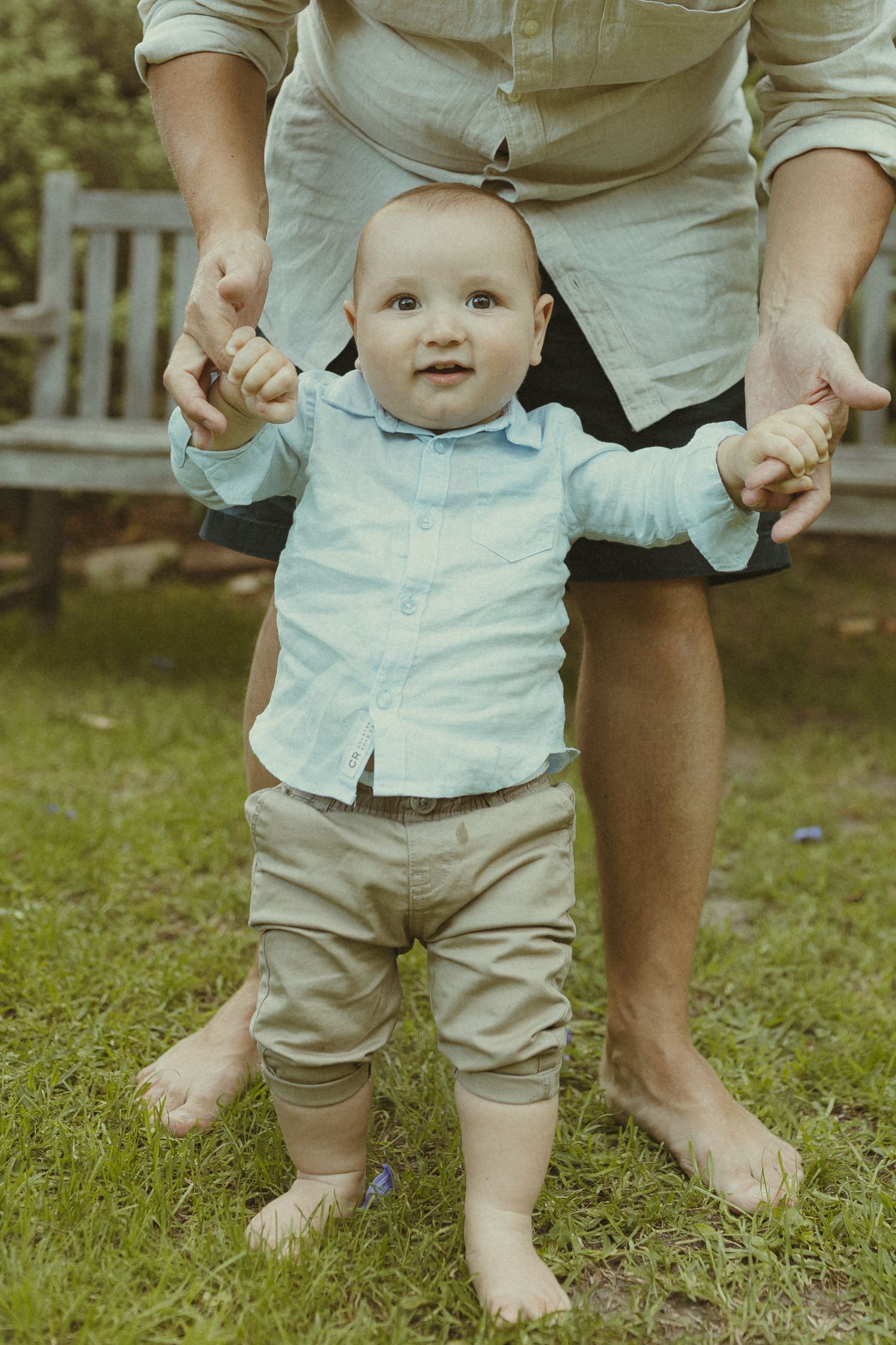 A toddler in a light blue shirt and khaki shorts stands barefoot on green grass during a sunny day.