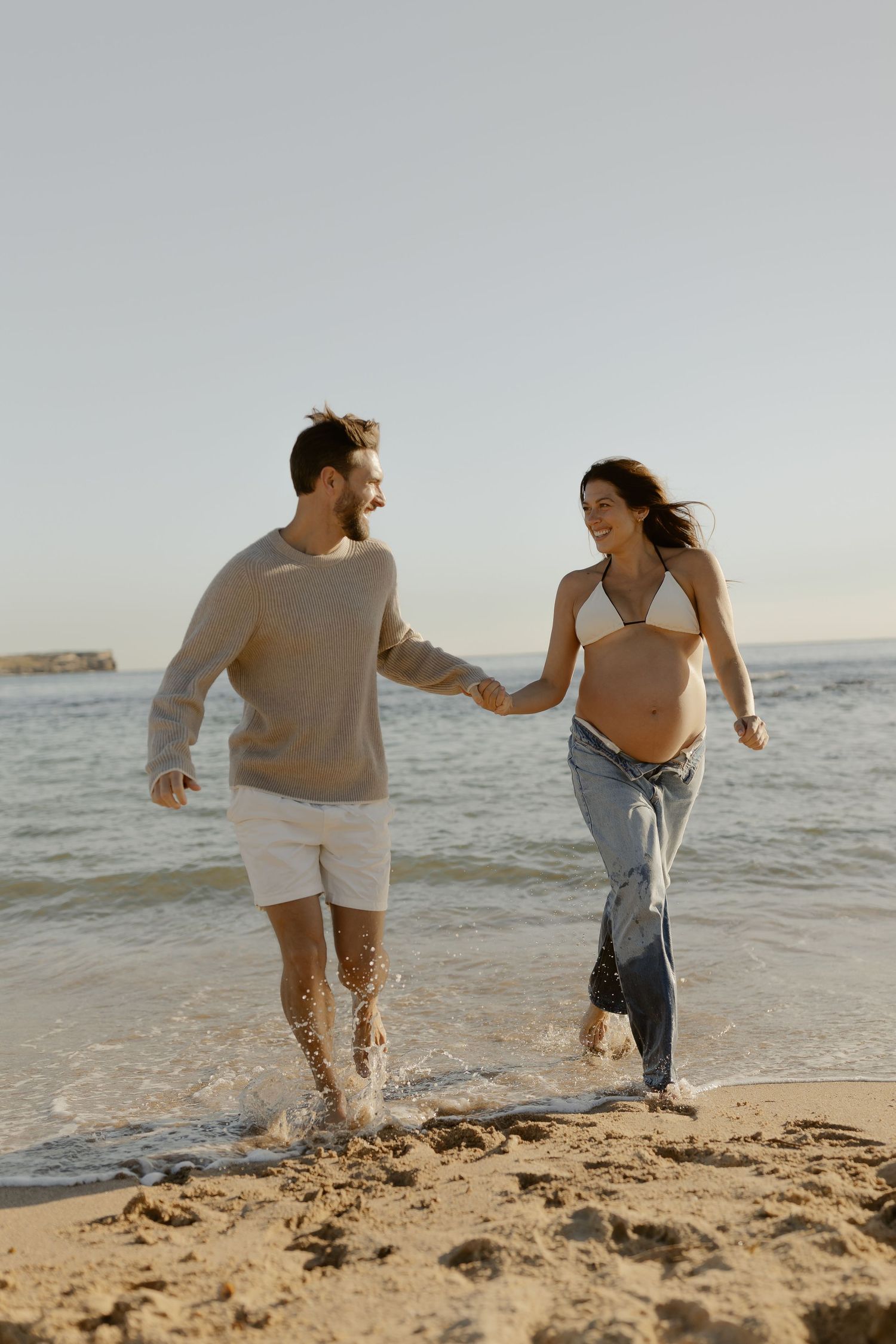 Couple running and splashing through shallow ocean water on sunny beach.