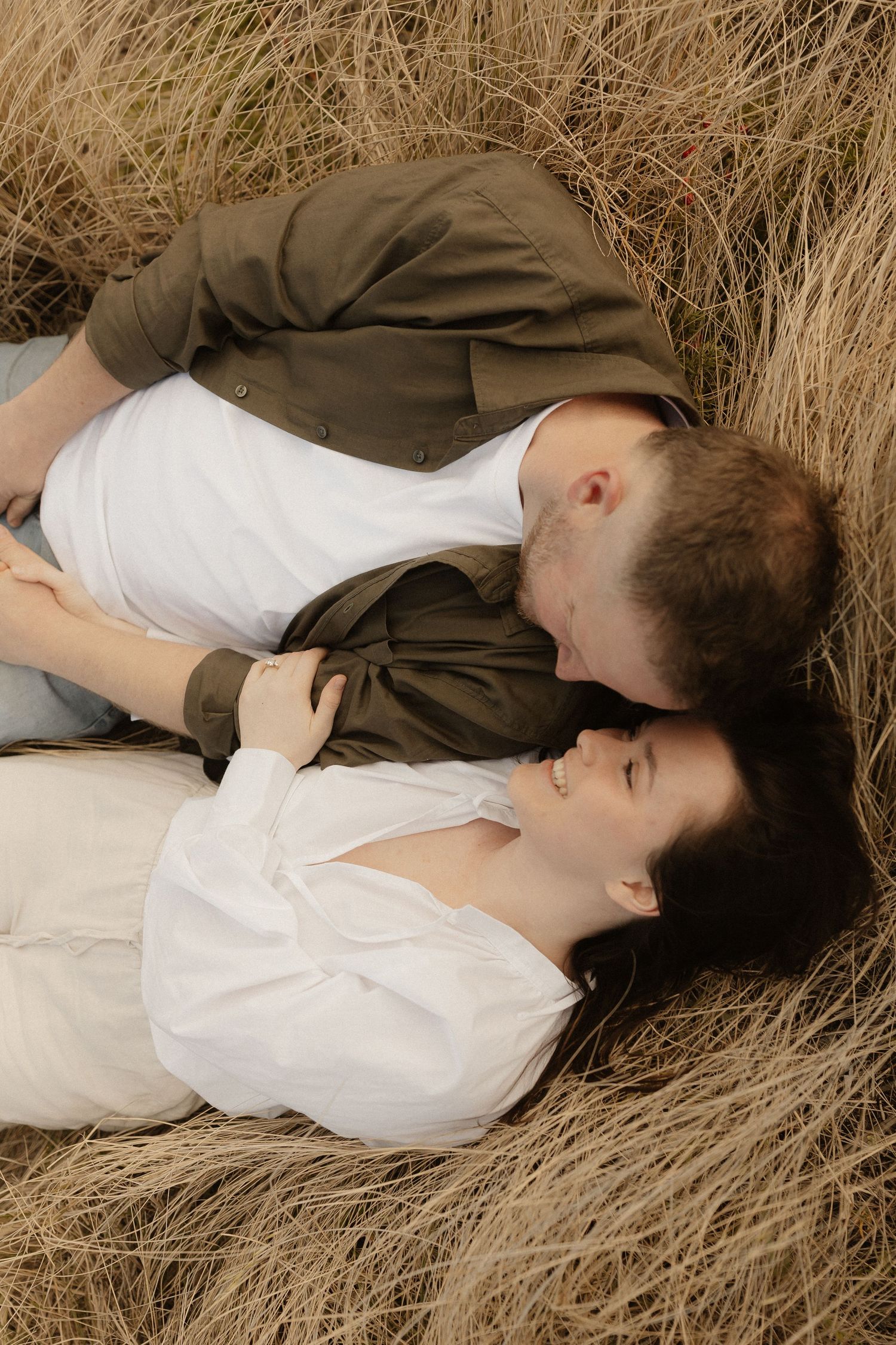 A couple lying together in tall brown grass creates an intimate outdoor portrait series.