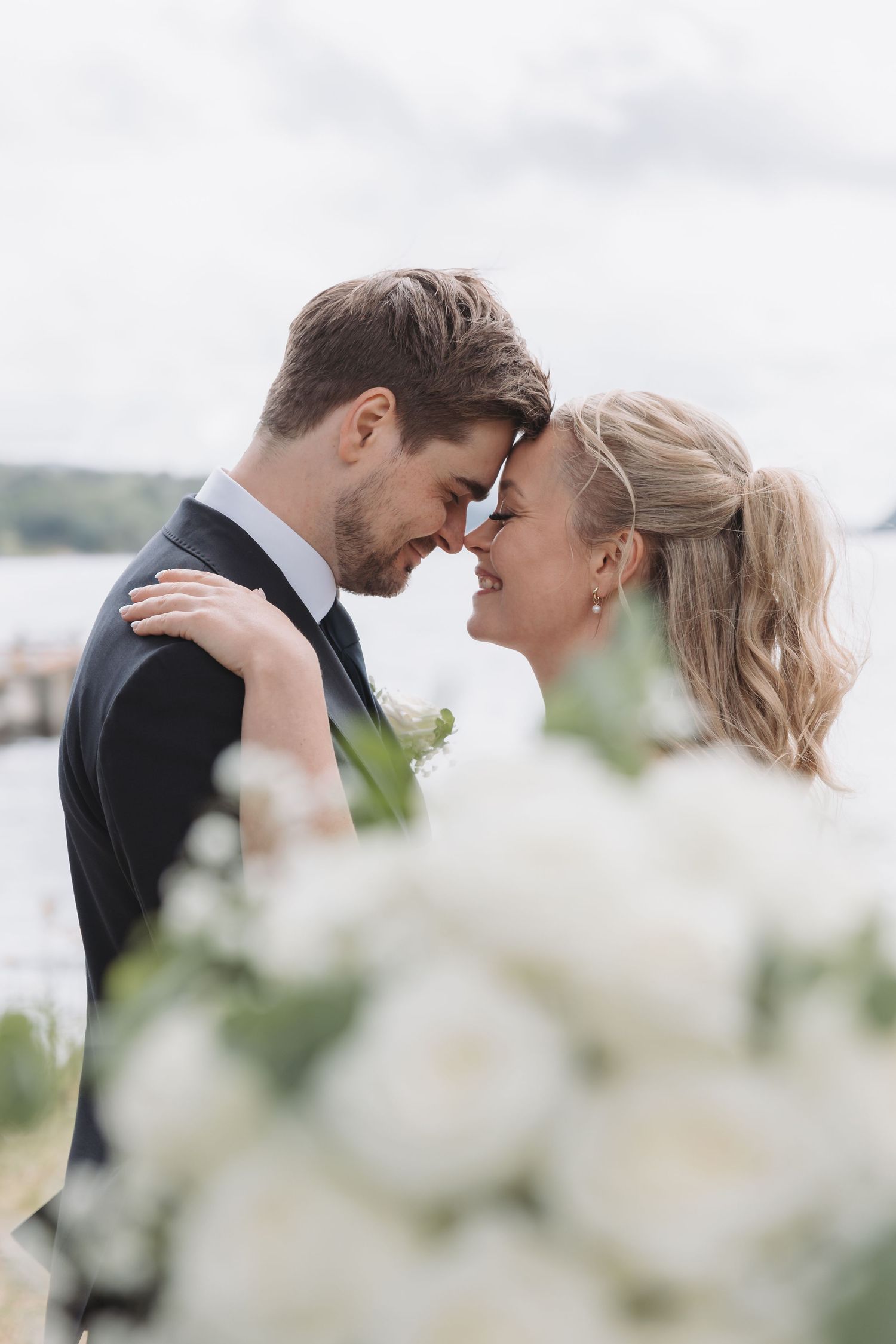 Bride and groom touching foreheads romantically with white flowers in foreground near waterfront.