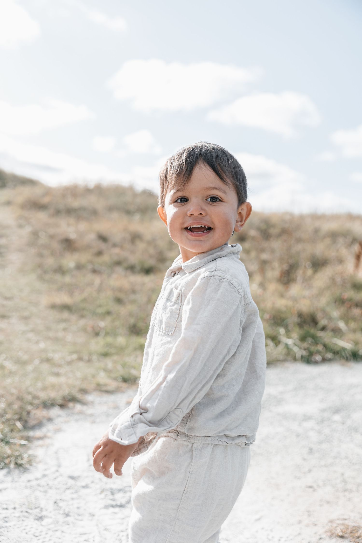Happy toddler in linen outfit smiling outdoors on a sunny dirt path with dry grass and blue sky background.