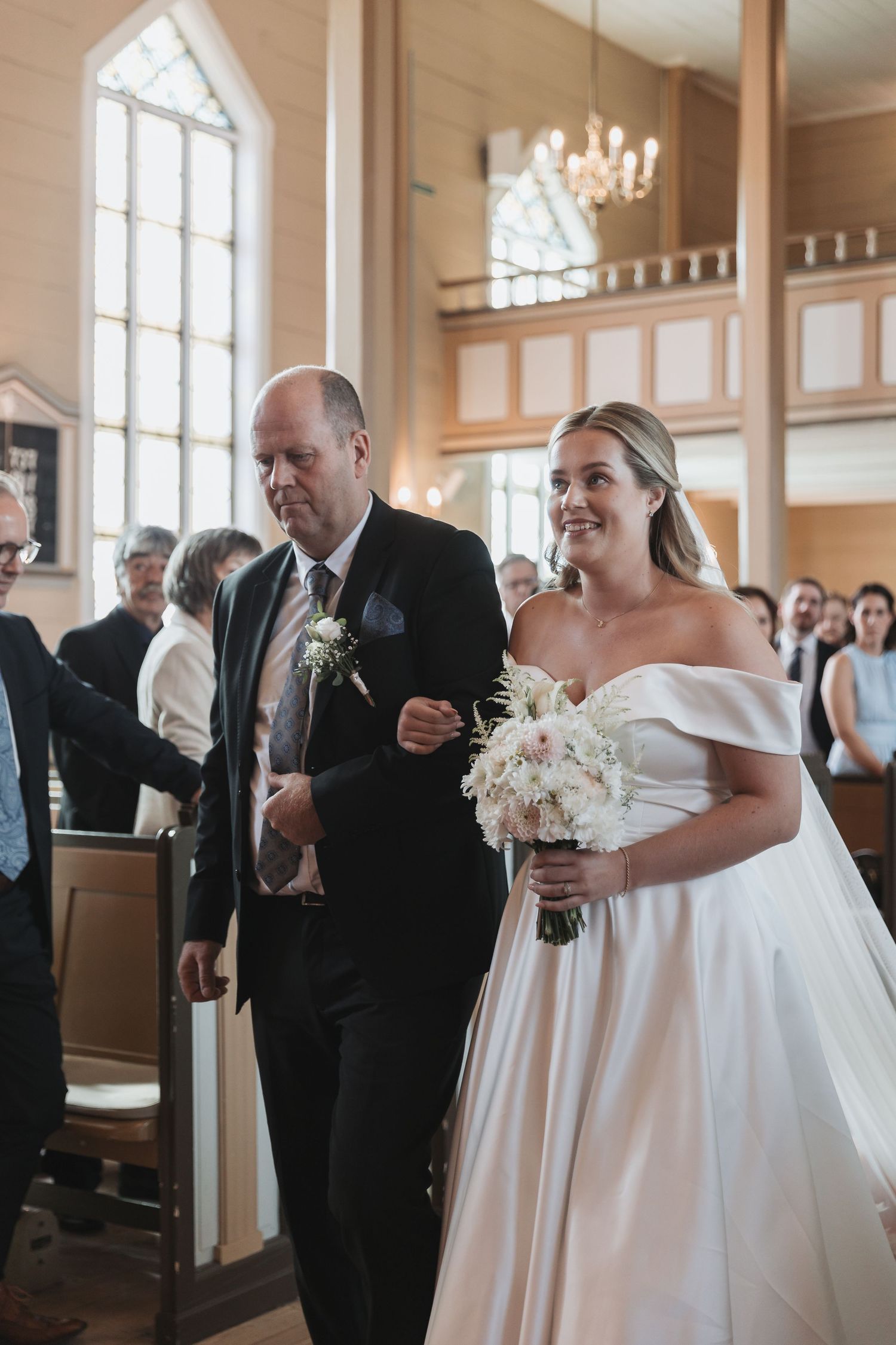 Bride in off-shoulder white gown holding bouquet walks down church aisle with escort during wedding ceremony.