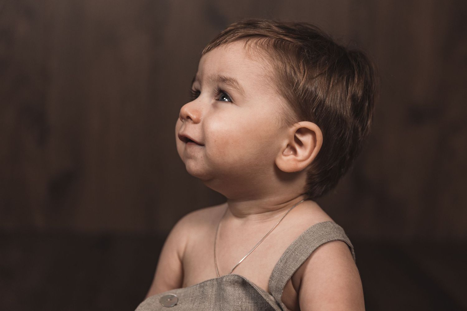 Young child in beige overalls gazes upward with wonder against dark wooden background.
