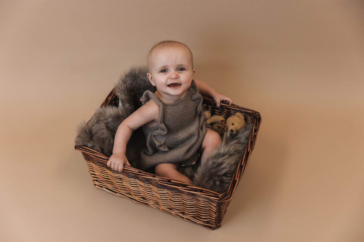 Happy baby sitting in wicker basket with cozy gray blanket against neutral beige backdrop during photo session.