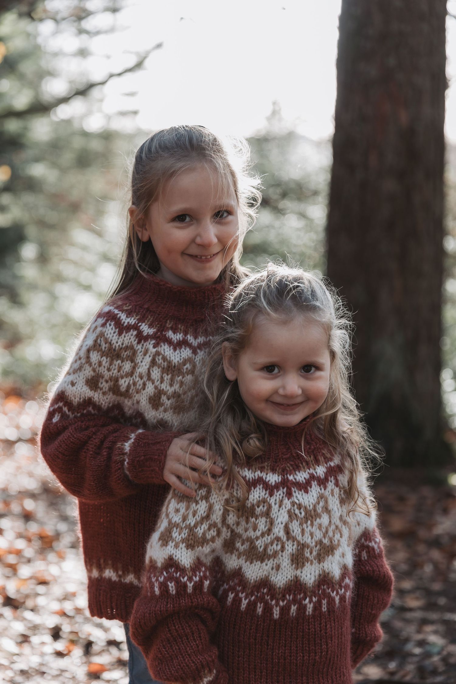 Two children wearing matching brown and white Fair Isle sweaters pose playfully in a forest setting.